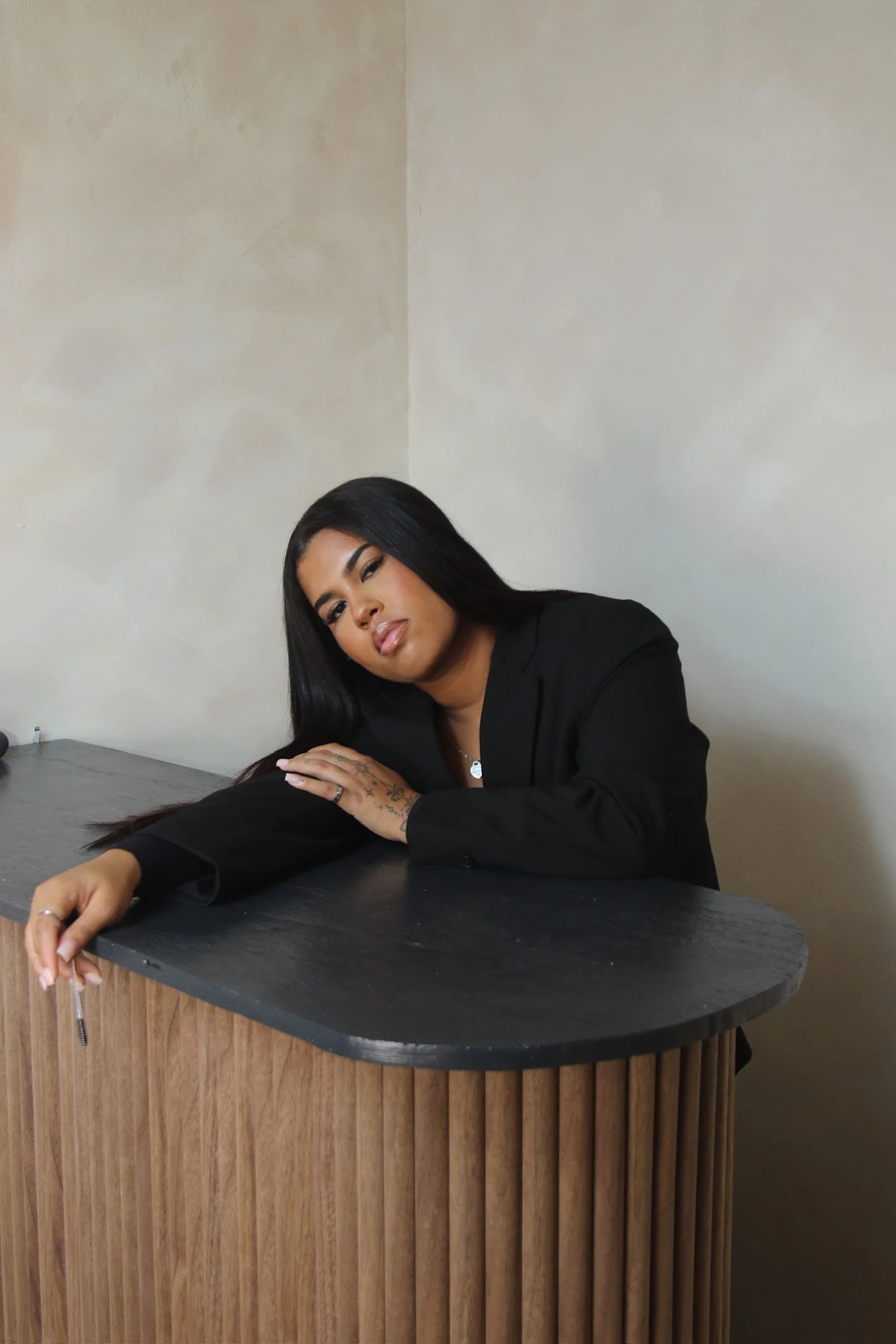 An Esthetician with long black hair, wearing a black blazer and touching a black desk with one arm, tilts her head slightly while looking at the camera inside a room with beige walls.