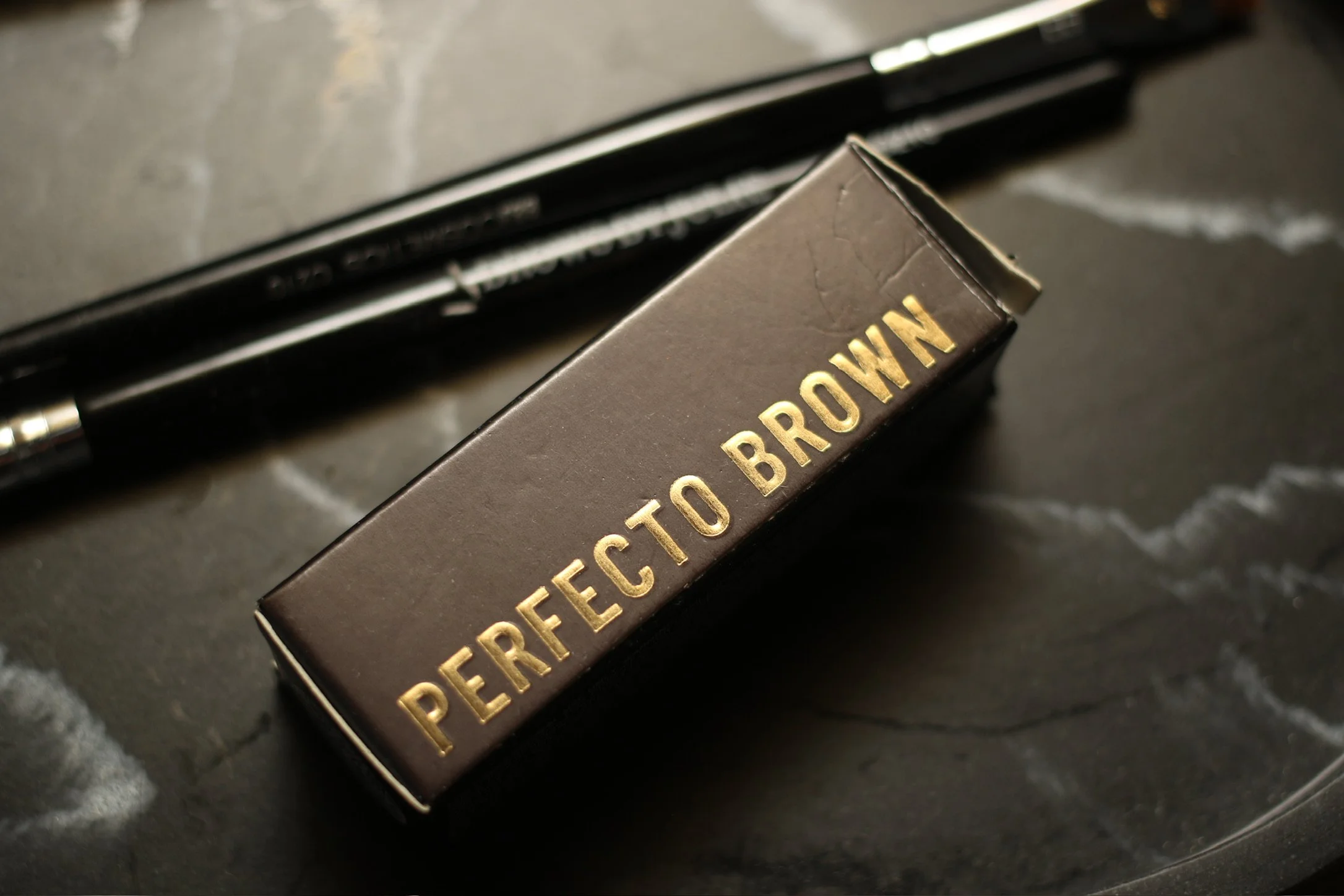 Close-up of a small black box with gold lettering that reads 'PERFECT TO BROWN' on a dark marble surface, with a black pen in the background.