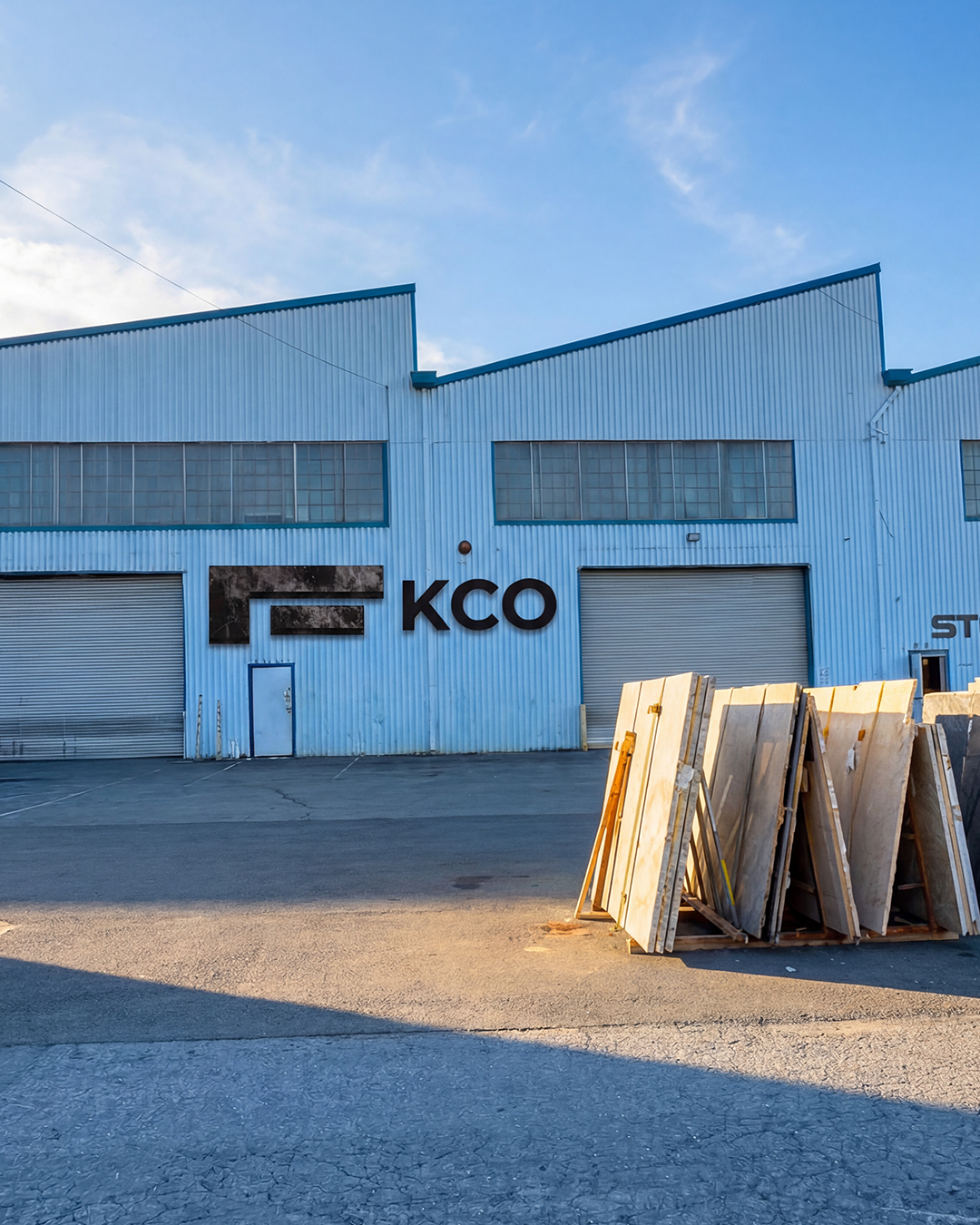 An industrial warehouse building with the letters 'KCO' on the front, a closed roll-up door, a smaller door, and a stack of wooden pallets outside under a blue sky.