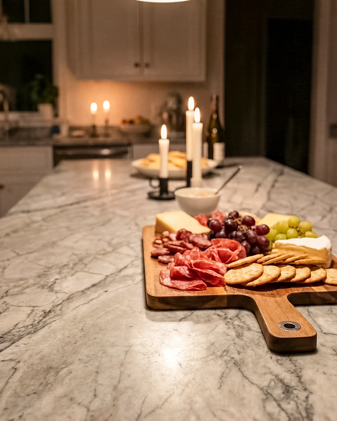 Charcuterie board with grapes, cheese, meat, crackers, and dip on kitchen marble countertop, lit by candles in the background.