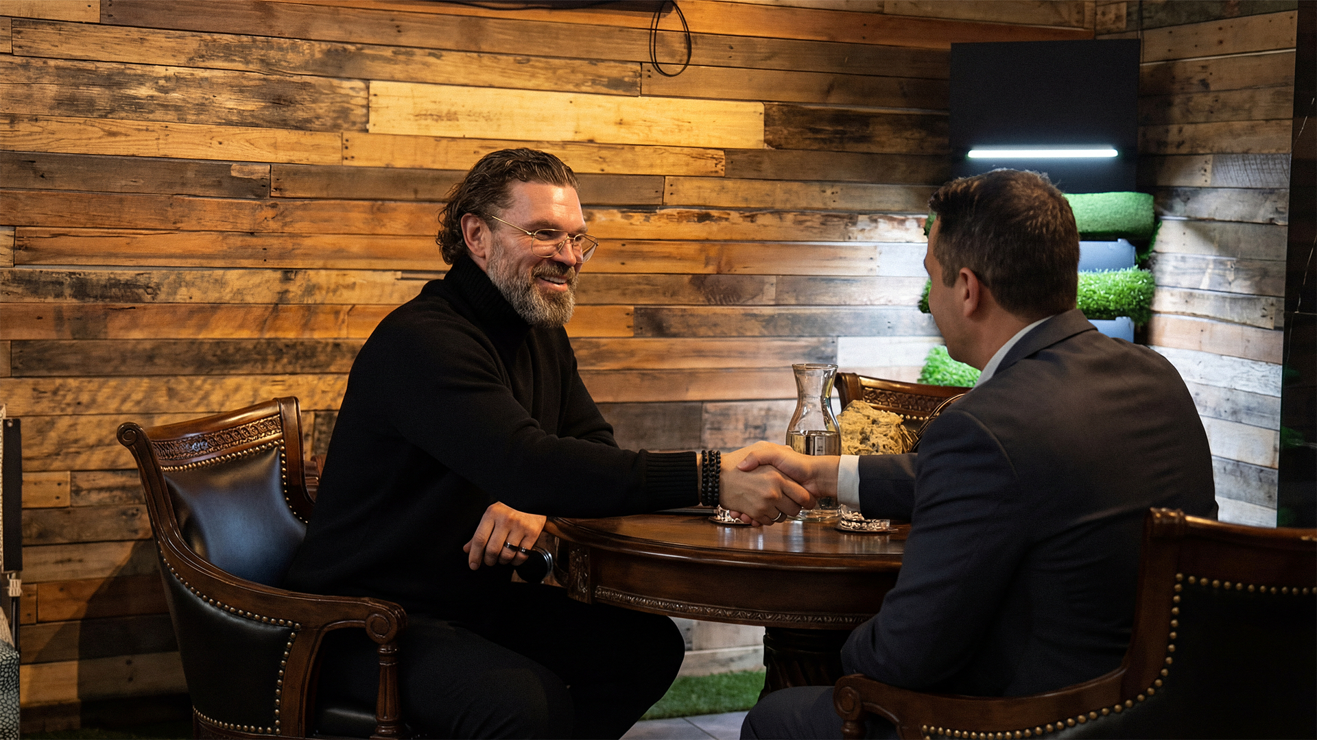 Two men shaking hands across a table in a warmly lit room with wood-paneled walls, one with gray hair and glasses, the other with dark hair, both dressed in dark clothing.