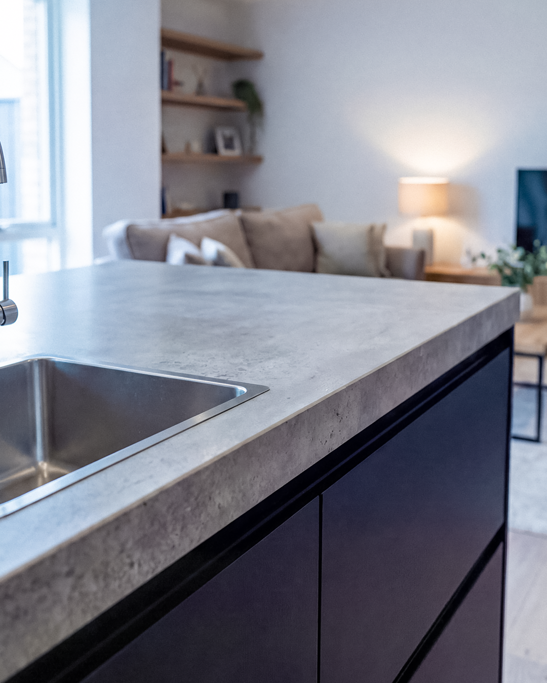 Close-up of a kitchen countertop with a built-in sink, with a living room visible in the background featuring a sofa, lamp, and shelves.