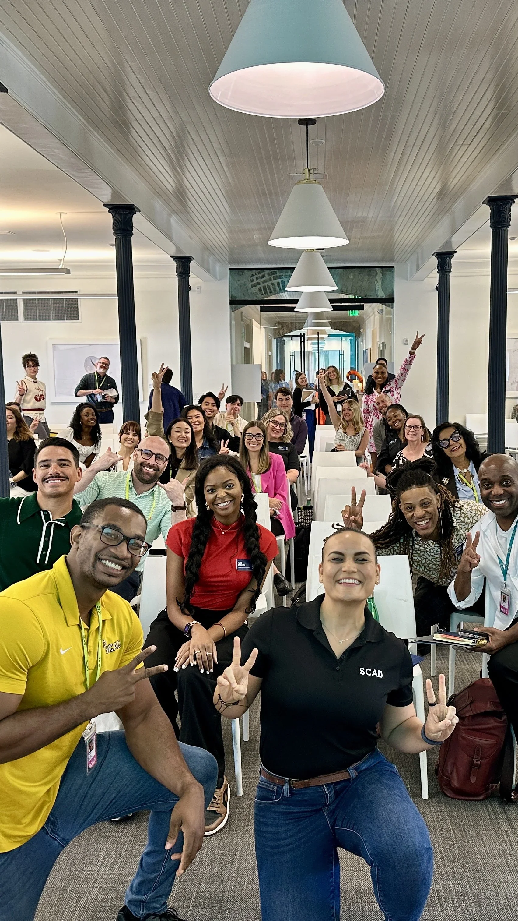 A woman with shoulder-length dark hair, smiling, wearing a purple coat and a black turtleneck, standing in front of a blurred window background.