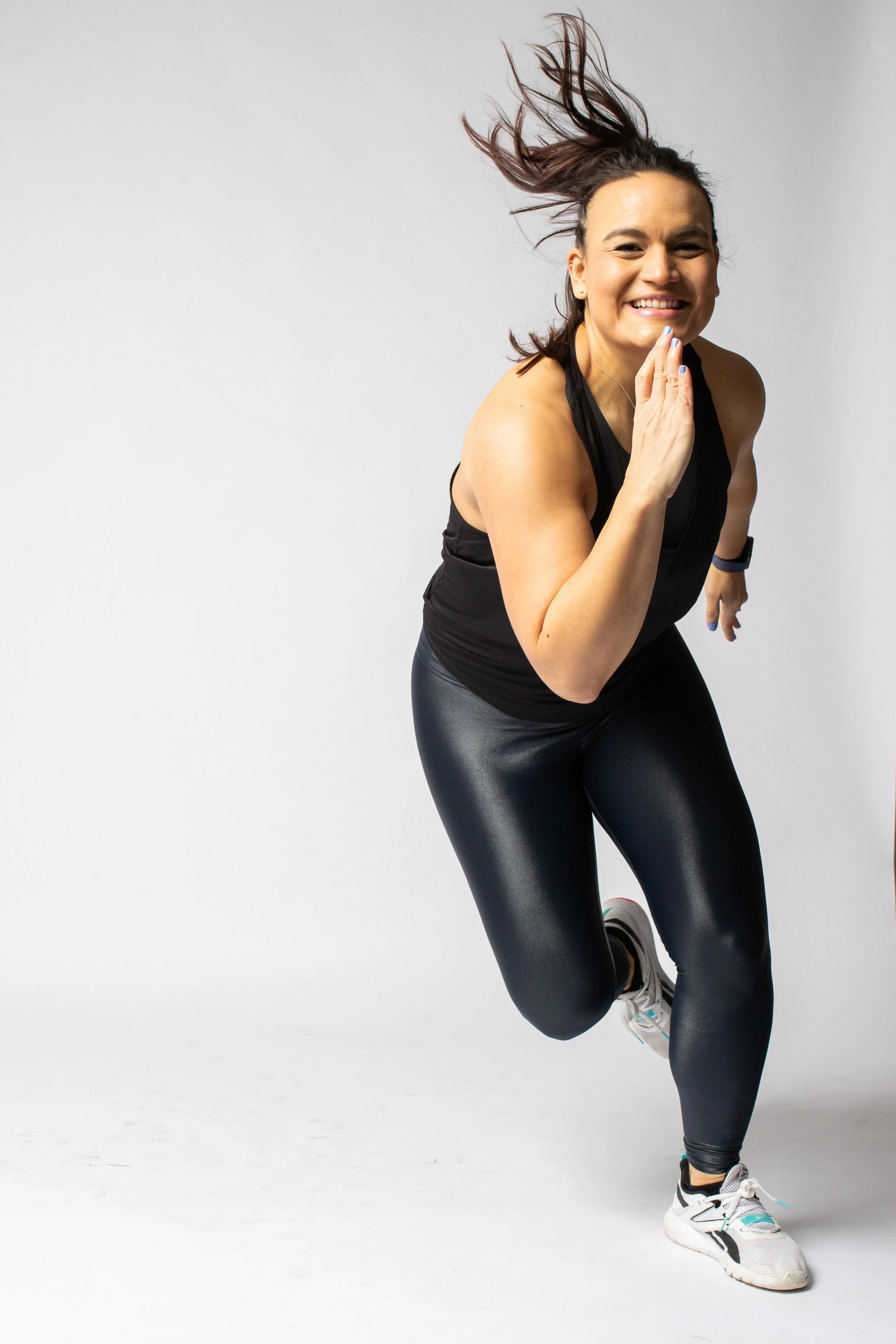 A woman dressed in athletic wear, smiling and running towards the camera against a plain background.
