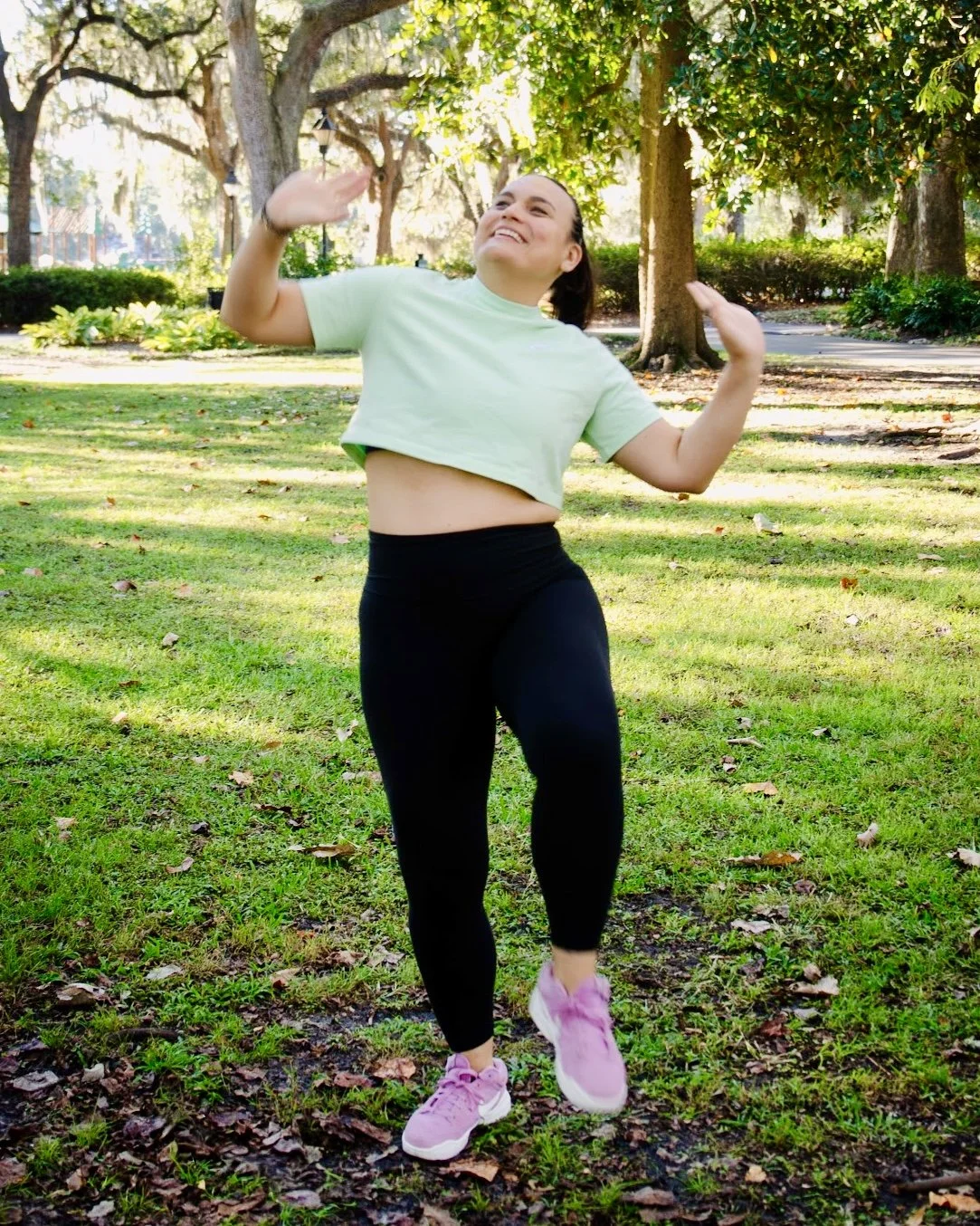 A woman in a green cropped t-shirt and black leggings dancing outdoors in a park with trees and grass.