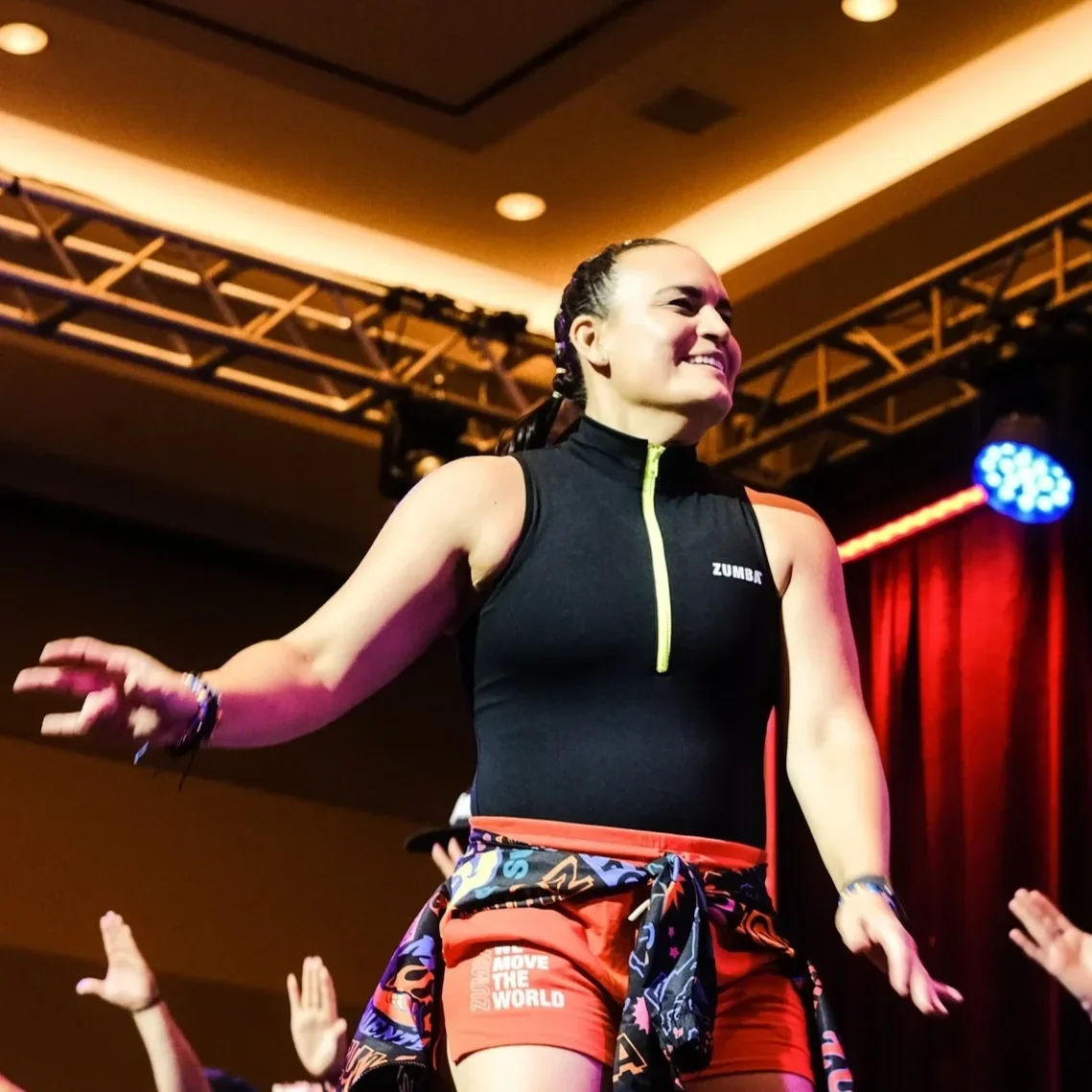 A woman smiling and joyfully jumping in a colorful studio with pink and blue lighting, wearing a black mesh top with 'ZUMBA' written on it and pink pants.