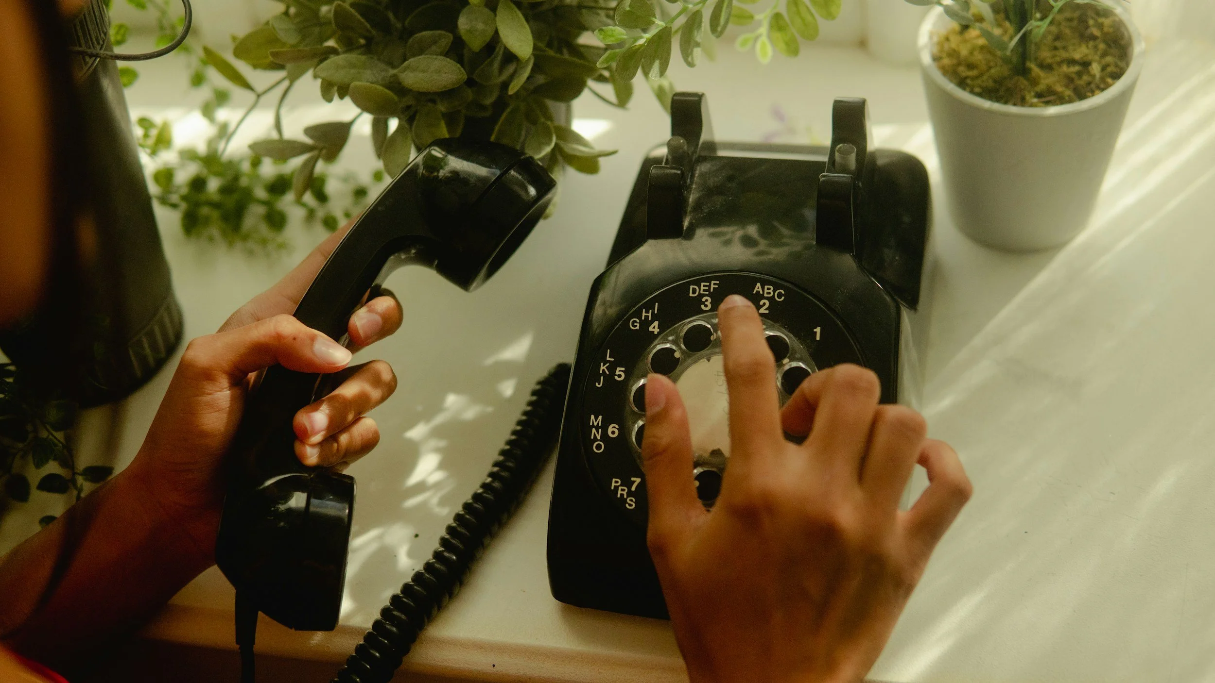Person dialing a number on a vintage black rotary phone with a dial having letters and numbers, placed on a table with potted plants nearby.