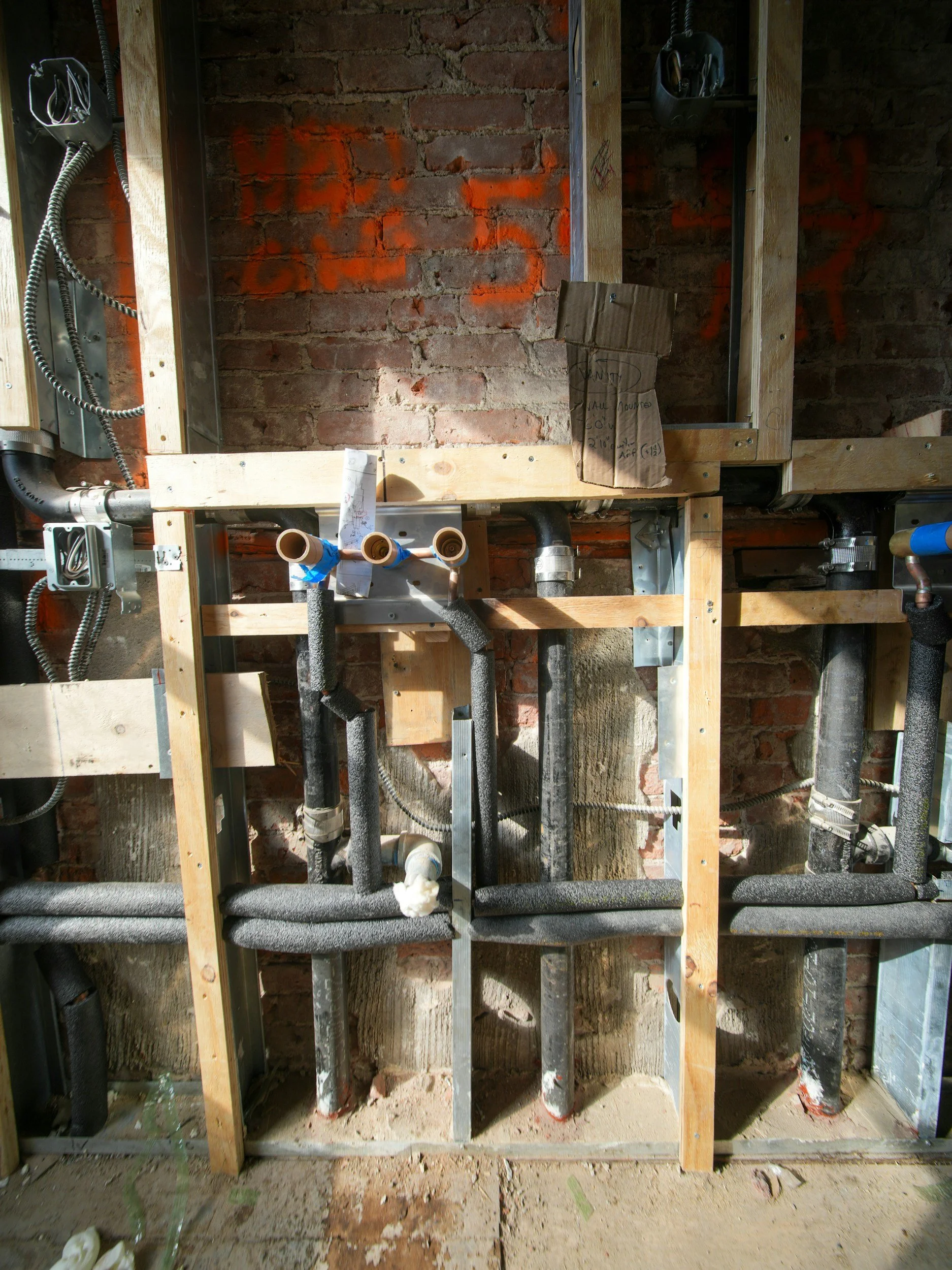 Construction site showing plumbing pipes and electrical wiring within a wooden frame against a brick wall.