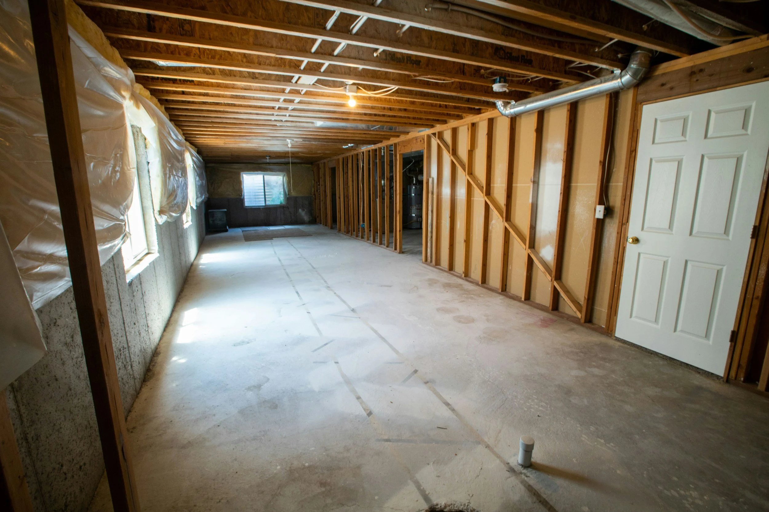 Unfinished basement with exposed wooden ceiling beams, insulation, and a concrete floor. Windows covered with plastic, with some natural light coming through. A white door on the right side, and pipes and ductwork are visible.