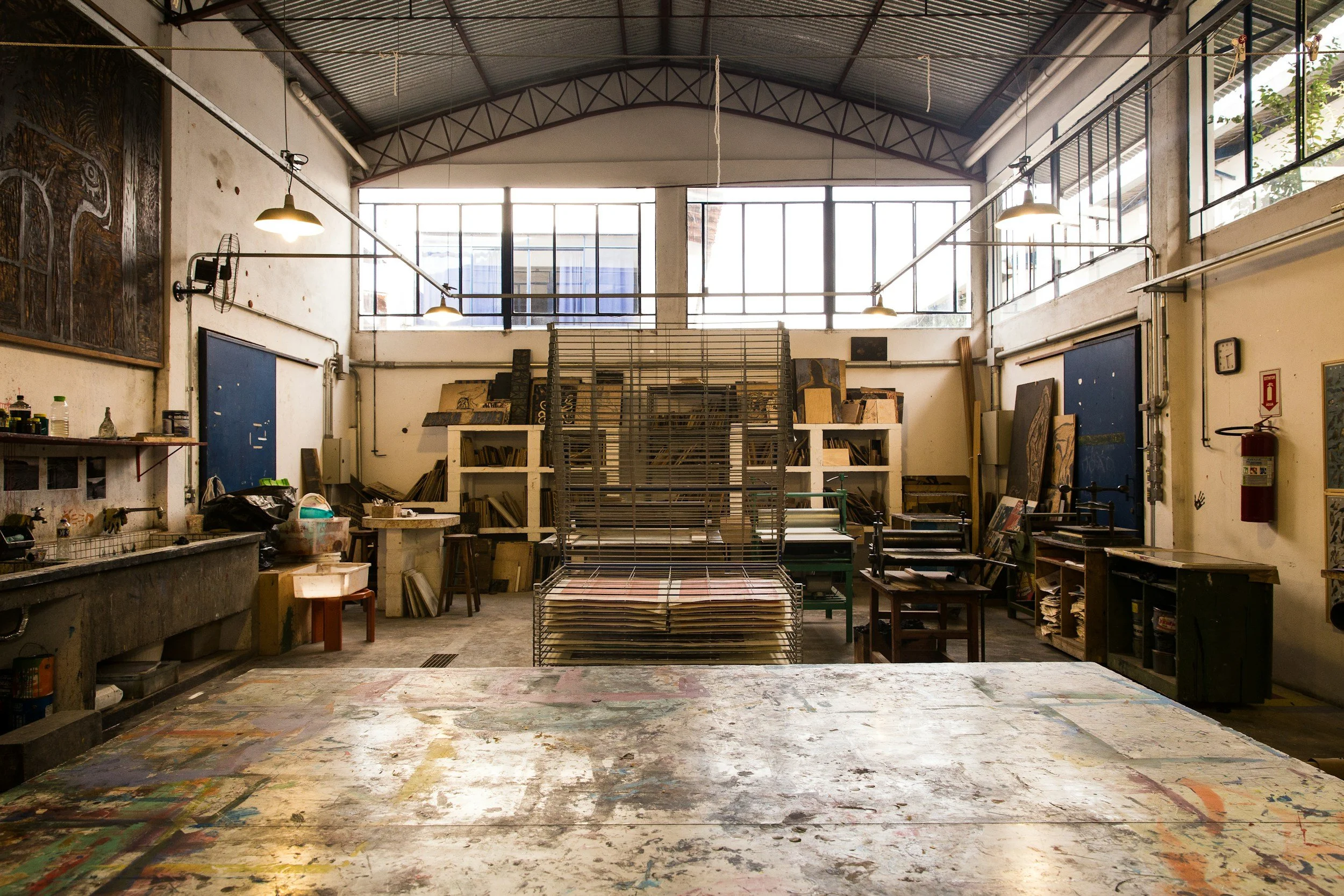 Art studio with large work table, shelves filled with artwork and supplies, paintings leaning against walls, overhead lighting, and a high ceiling with windows.
