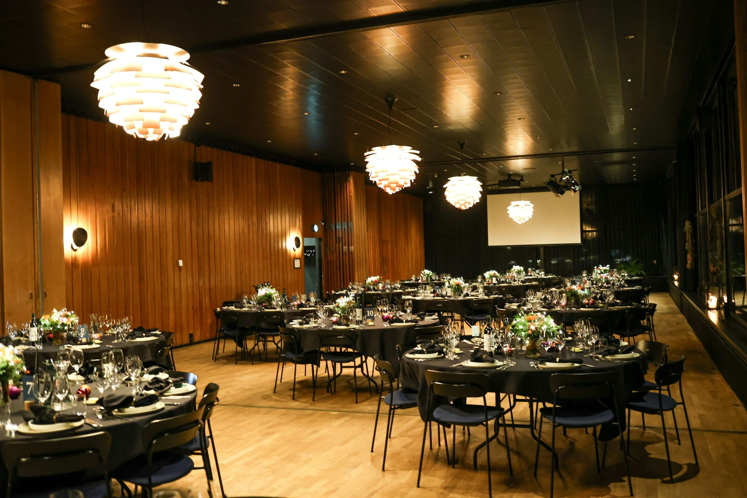 An elegantly decorated banquet hall with round tables covered with black tablecloths, set with plates, silverware, wine glasses, and floral centerpieces. The room features wooden panel walls, modern hanging light fixtures, and a large projection screen at the front.