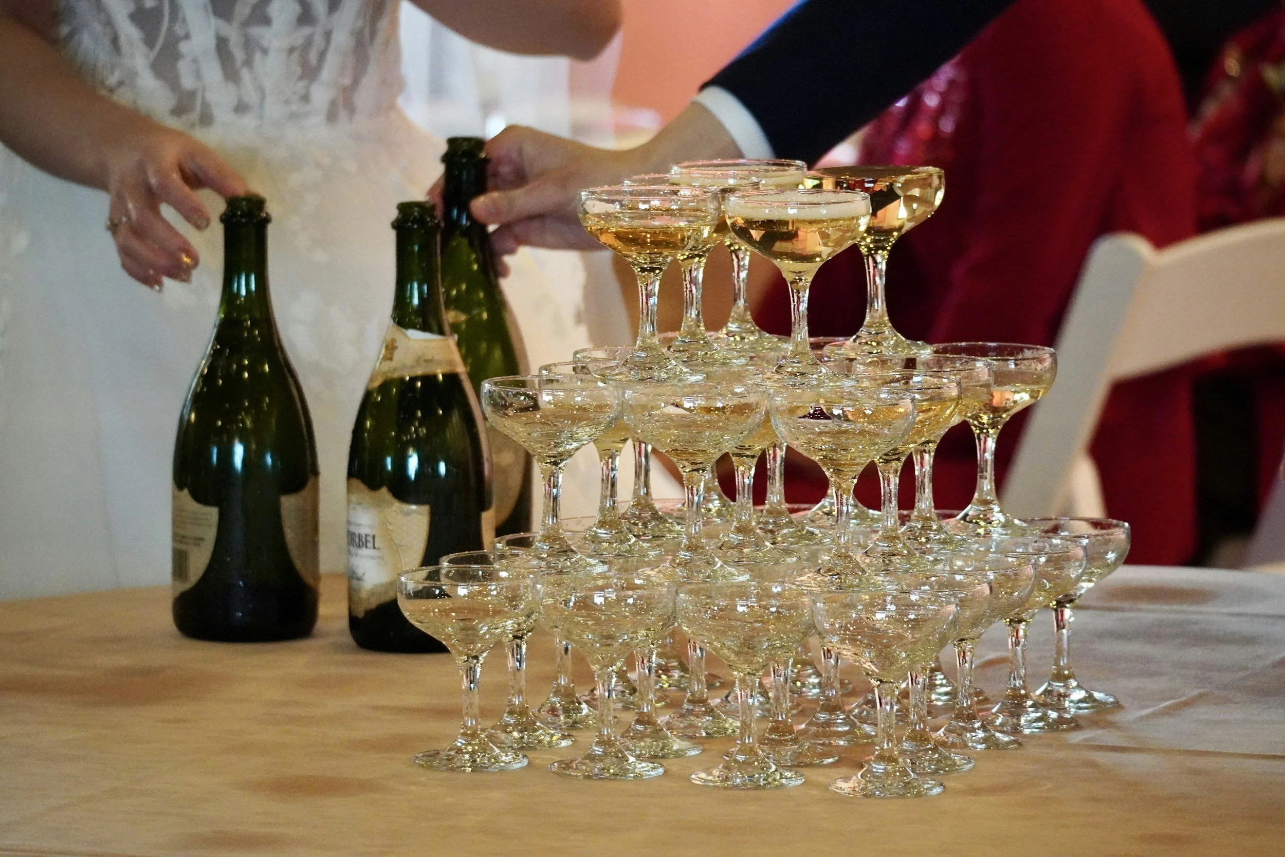 Champagne glasses stacked in a pyramid on a table with bottles of champagne in the background at a celebration or wedding.
