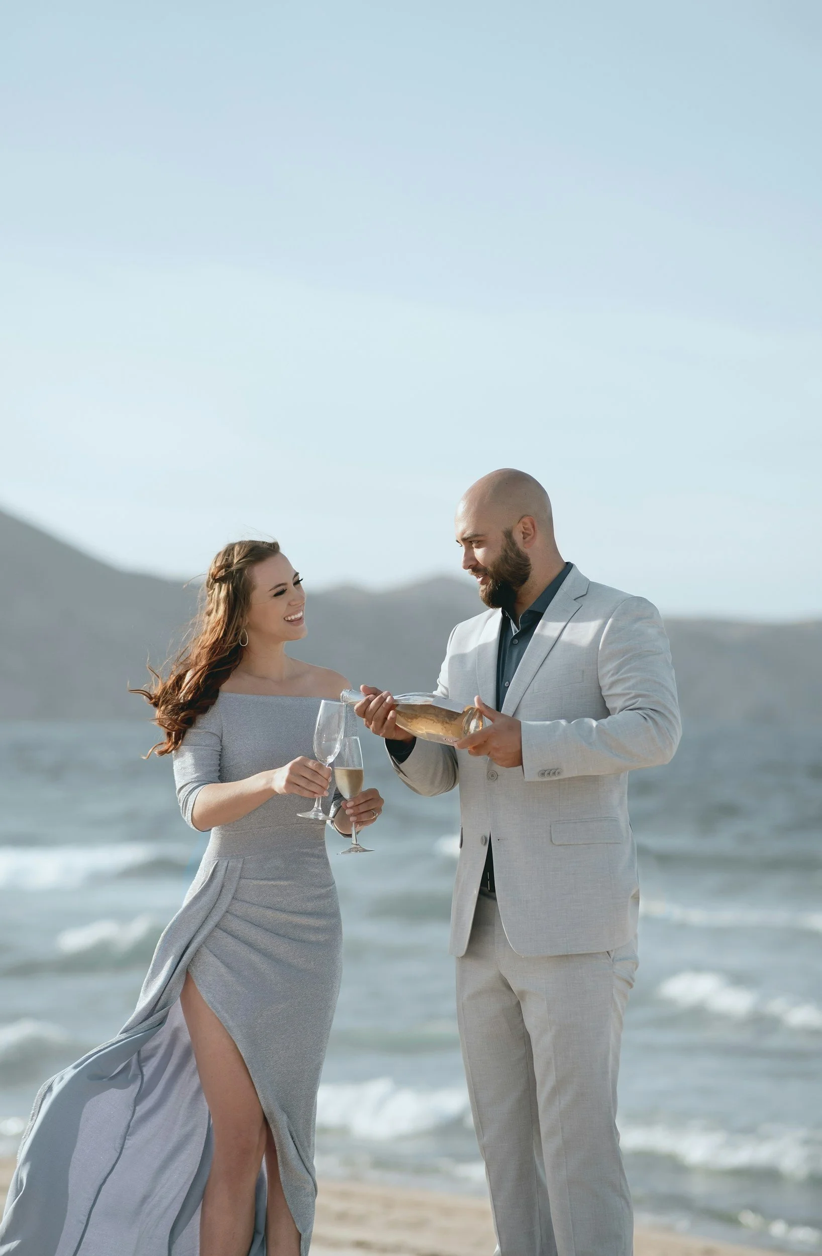 A man in a light gray suit offers a bottle of champagne to a woman in a light gray off-shoulder dress with a slit, both on a beach with the ocean and mountains in the background.