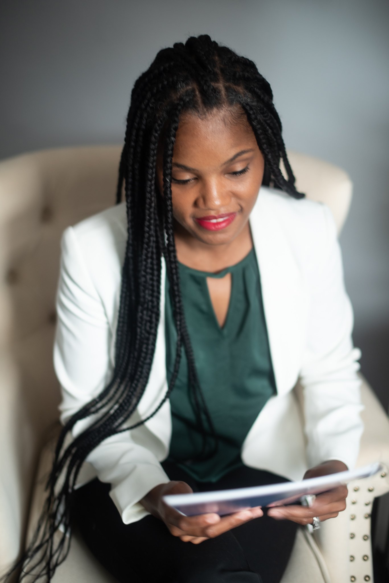 A woman with long braided hair, wearing a white blazer and a dark green blouse, sitting on a beige armchair, looking at a tablet.