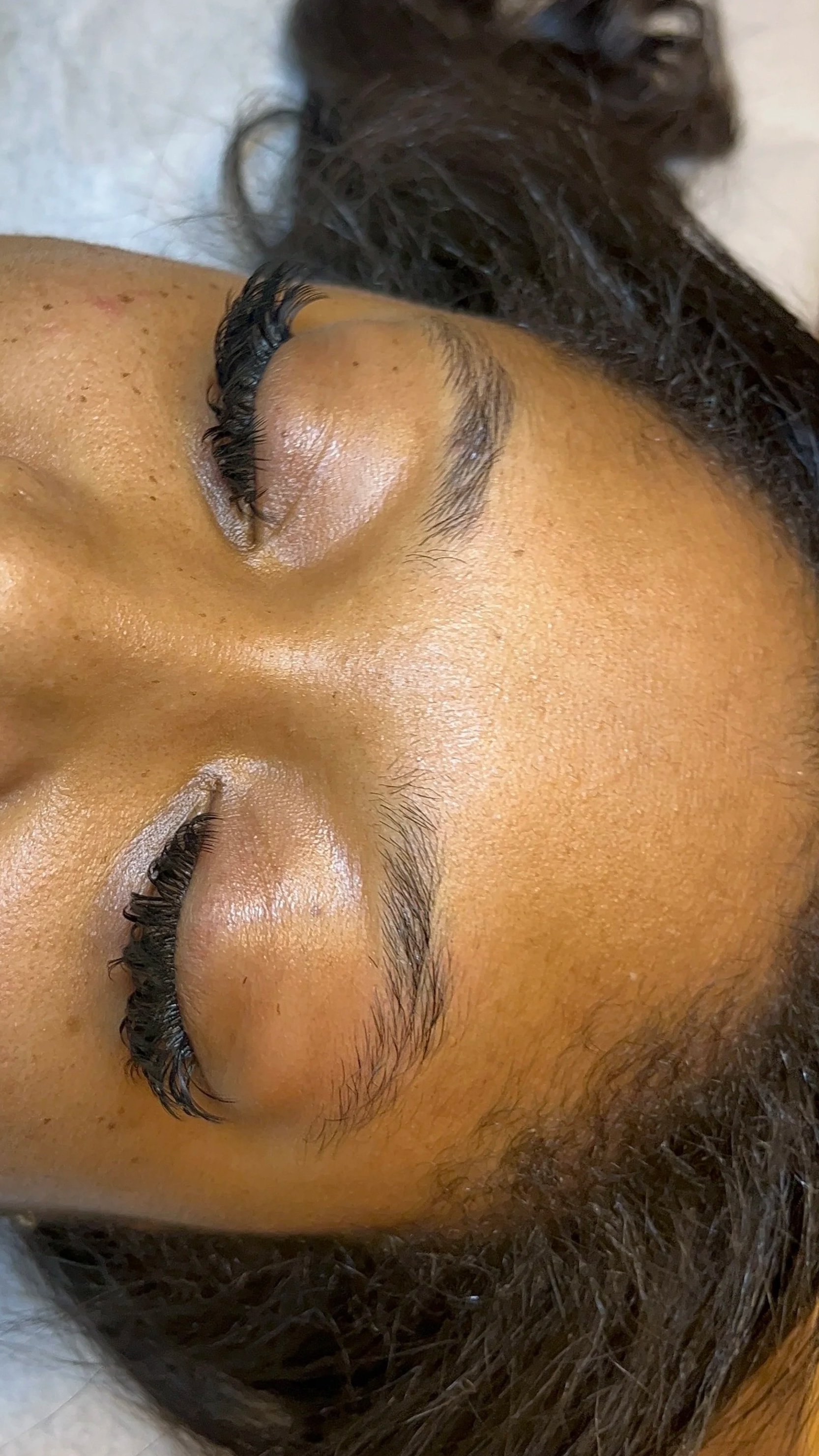 Close-up of a woman's face with eyes closed, showing her eyebrows, eyelash extensions, and skin texture.