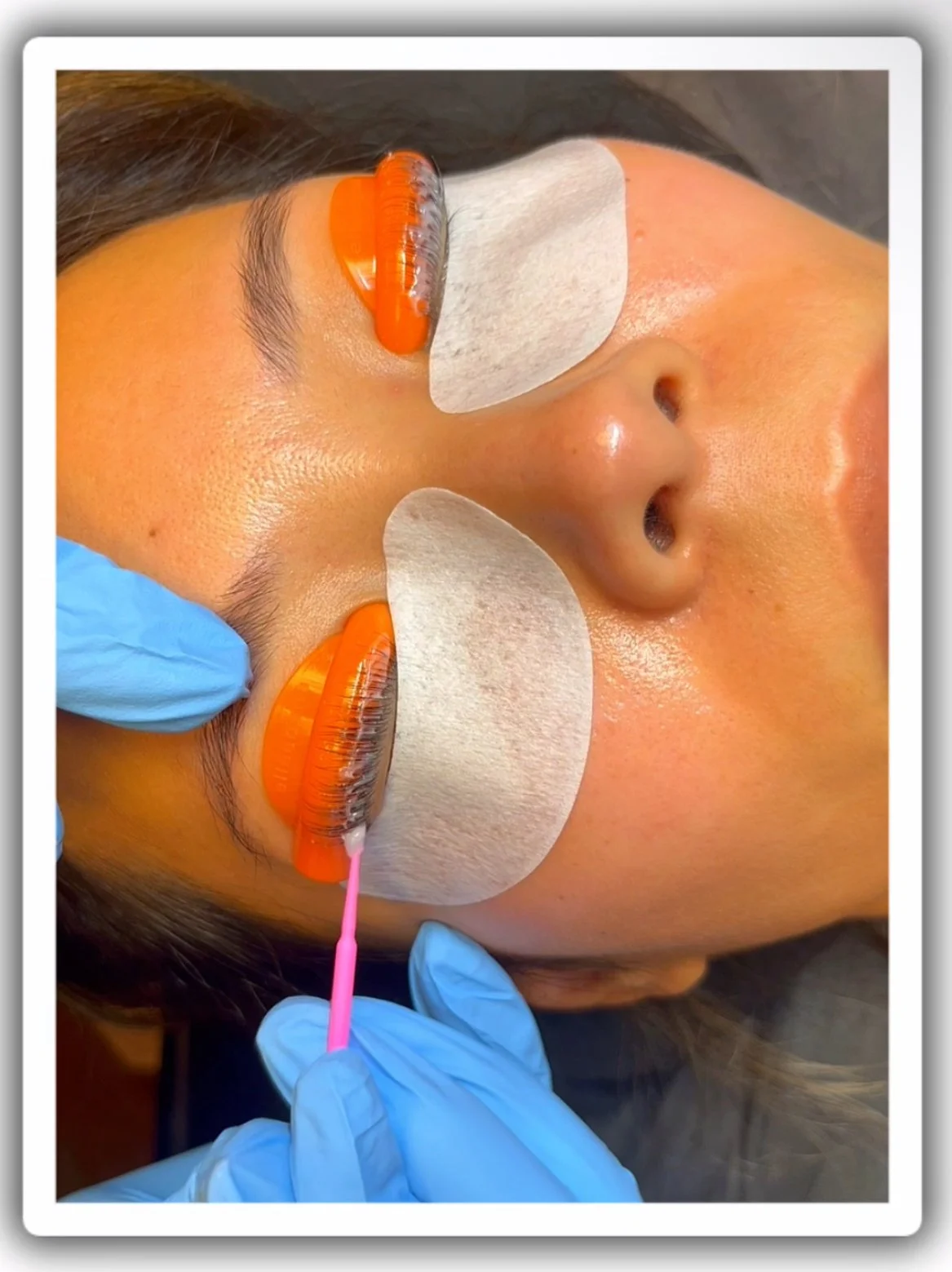 A woman receiving eyelash extension treatment with orange tape and cotton pads under her eyes, ellebana lash lift process