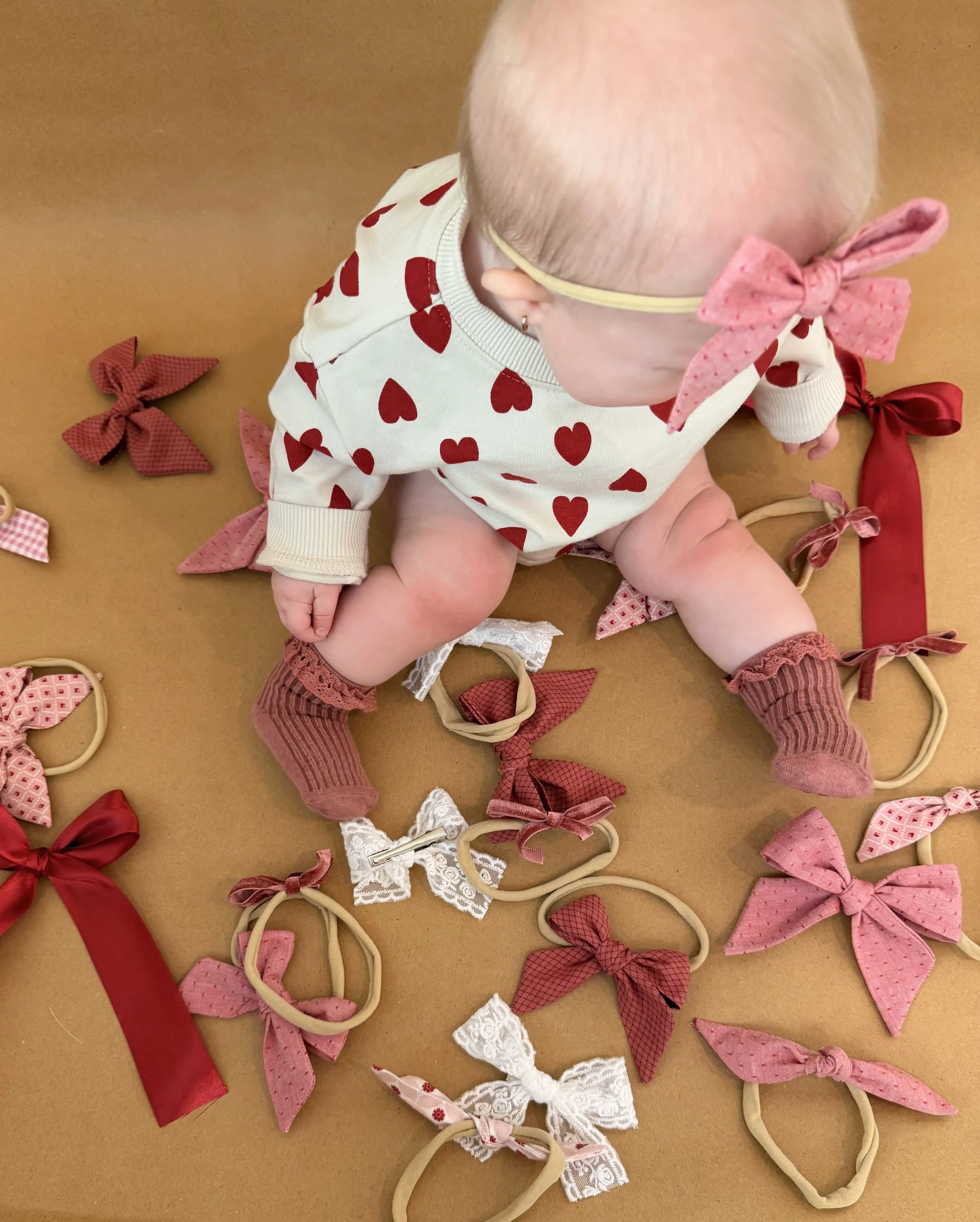 A baby girl wearing a white shirt with red hearts, pink socks, and a pink bow headband sitting on a brown surface surrounded by pink, red, and white bow-shaped hair ties.