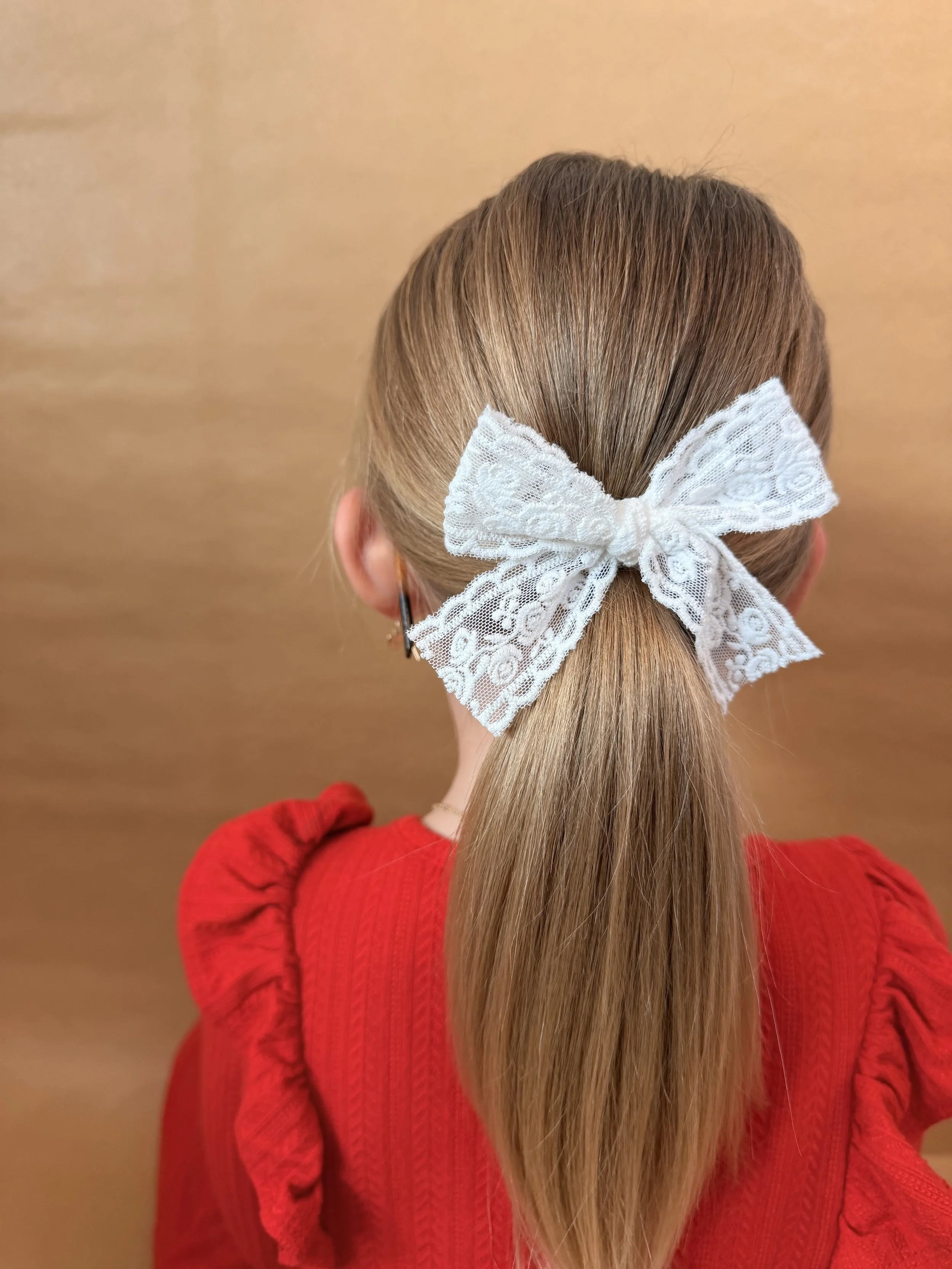 Back of a person's head with long, straight, light brown hair tied into a ponytail with a large white lace bow accessory. The person is wearing a red top with puffed sleeves, and a wooden background is visible.