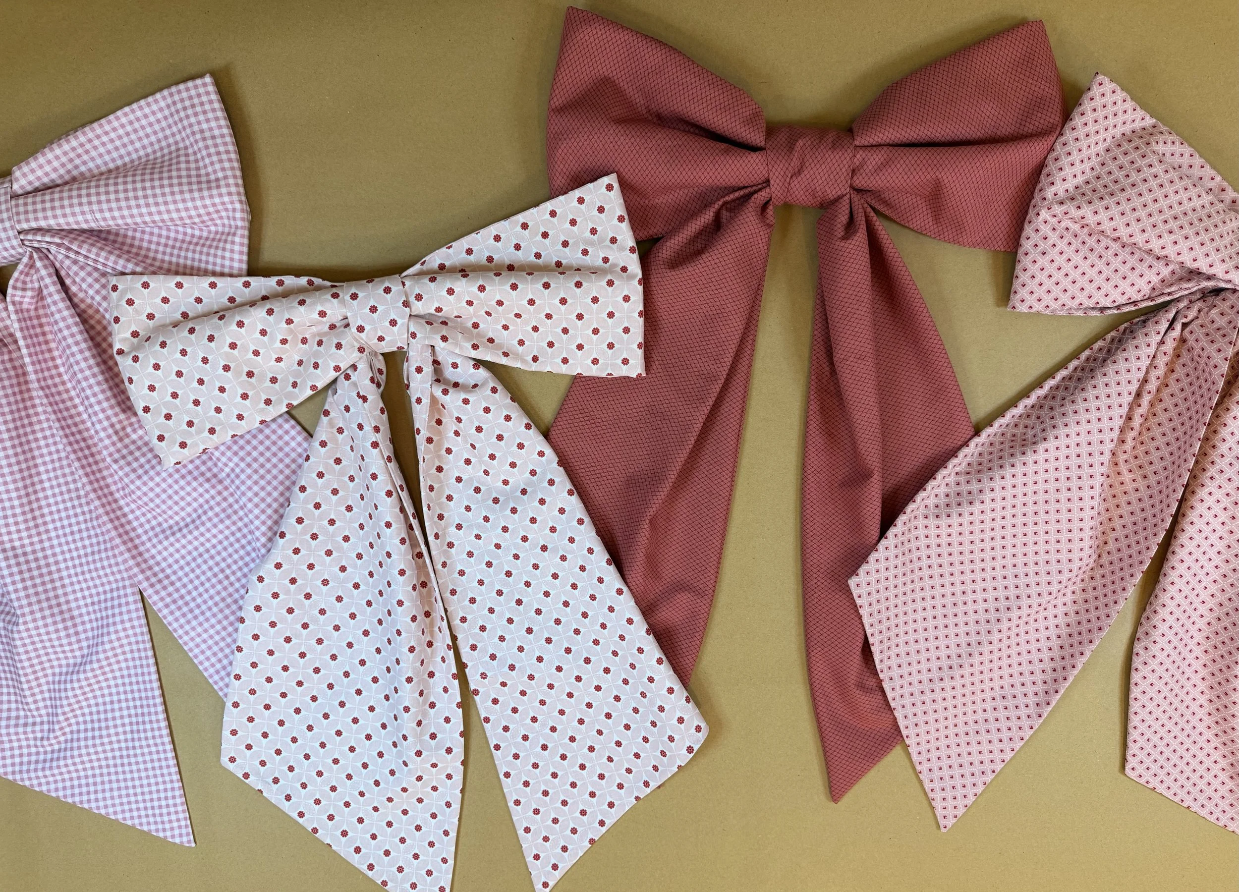Three large fabric bows in shades of pink and white, with patterns including checks, polka dots, and small geometric designs, arranged on a beige surface.