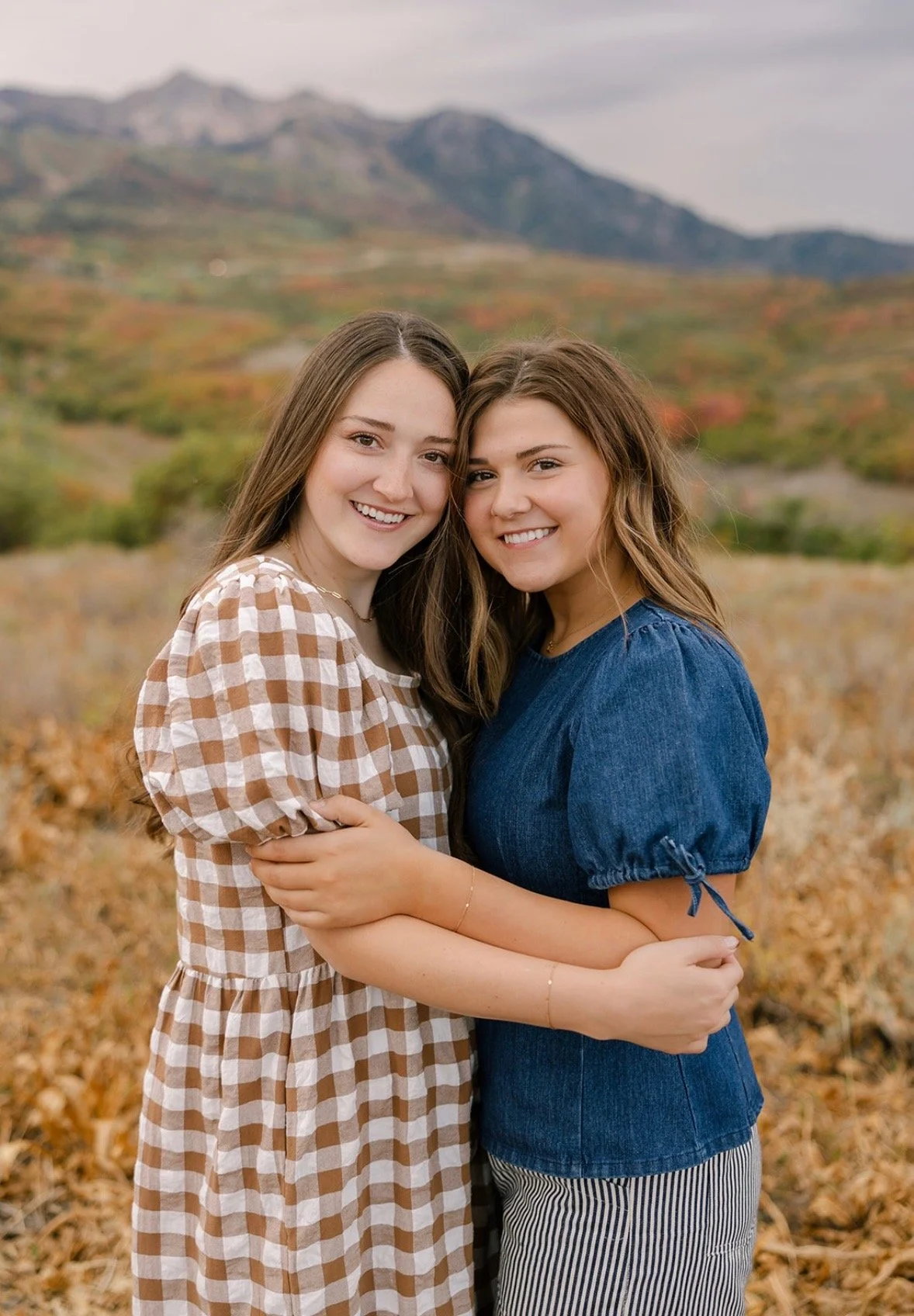 Two young women embracing outdoors with mountains in the background, wearing casual clothing, smiling at the camera.