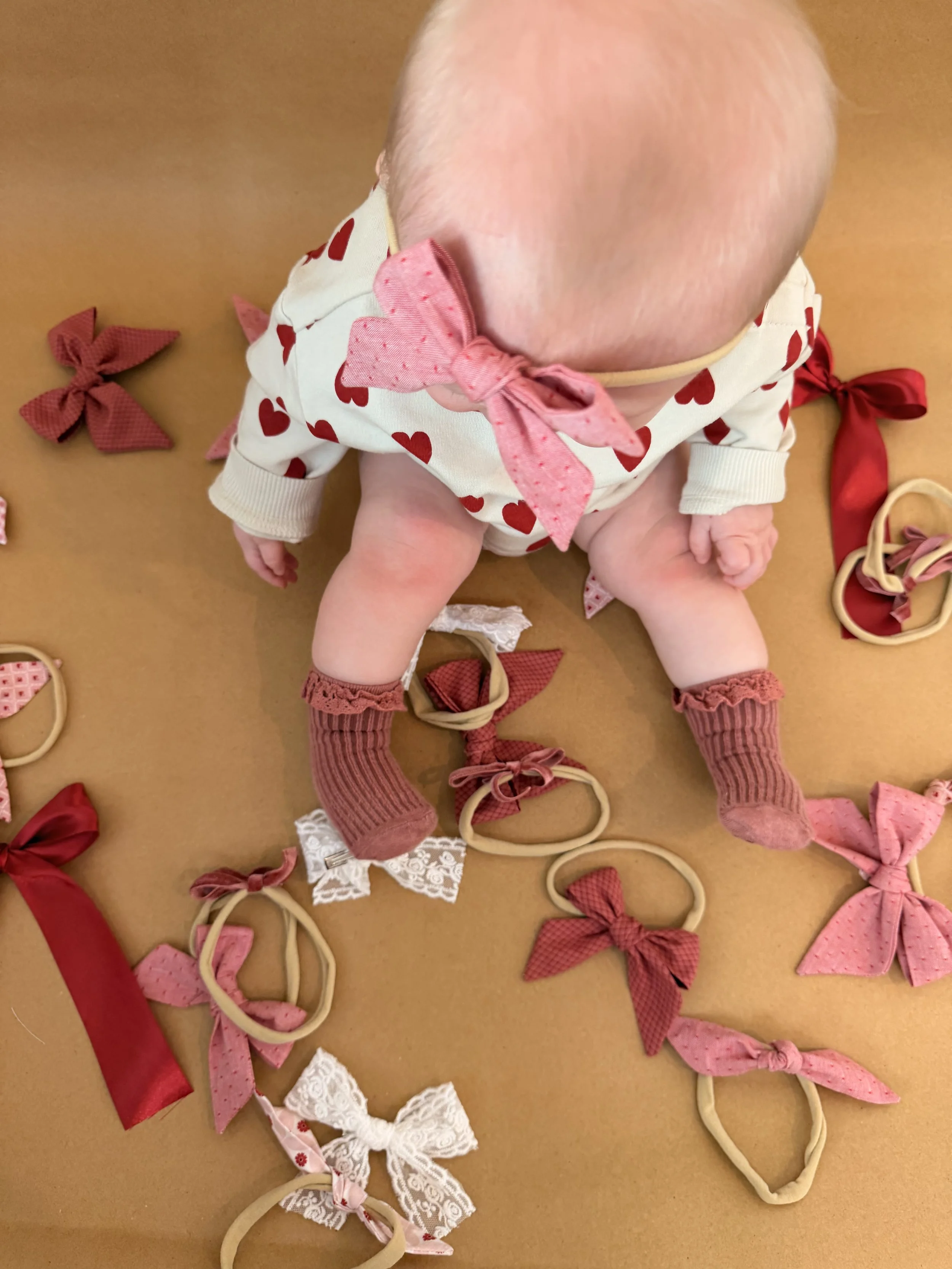 A baby sitting on a brown surface surrounded by pink, red, and cream-colored hair ties with bows.