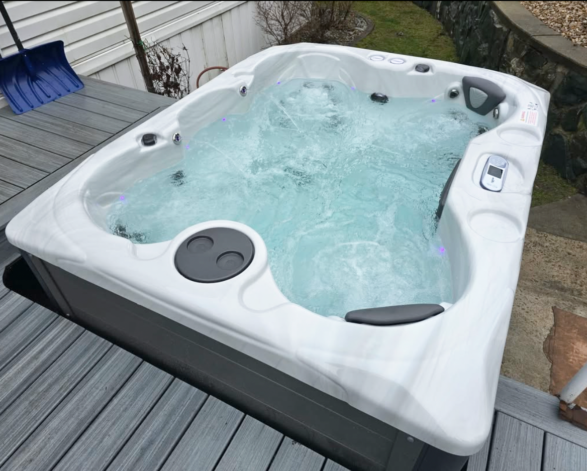 An outdoor hot tub with bubbling water on a wooden deck, surrounded by a stone wall and part of a yard.