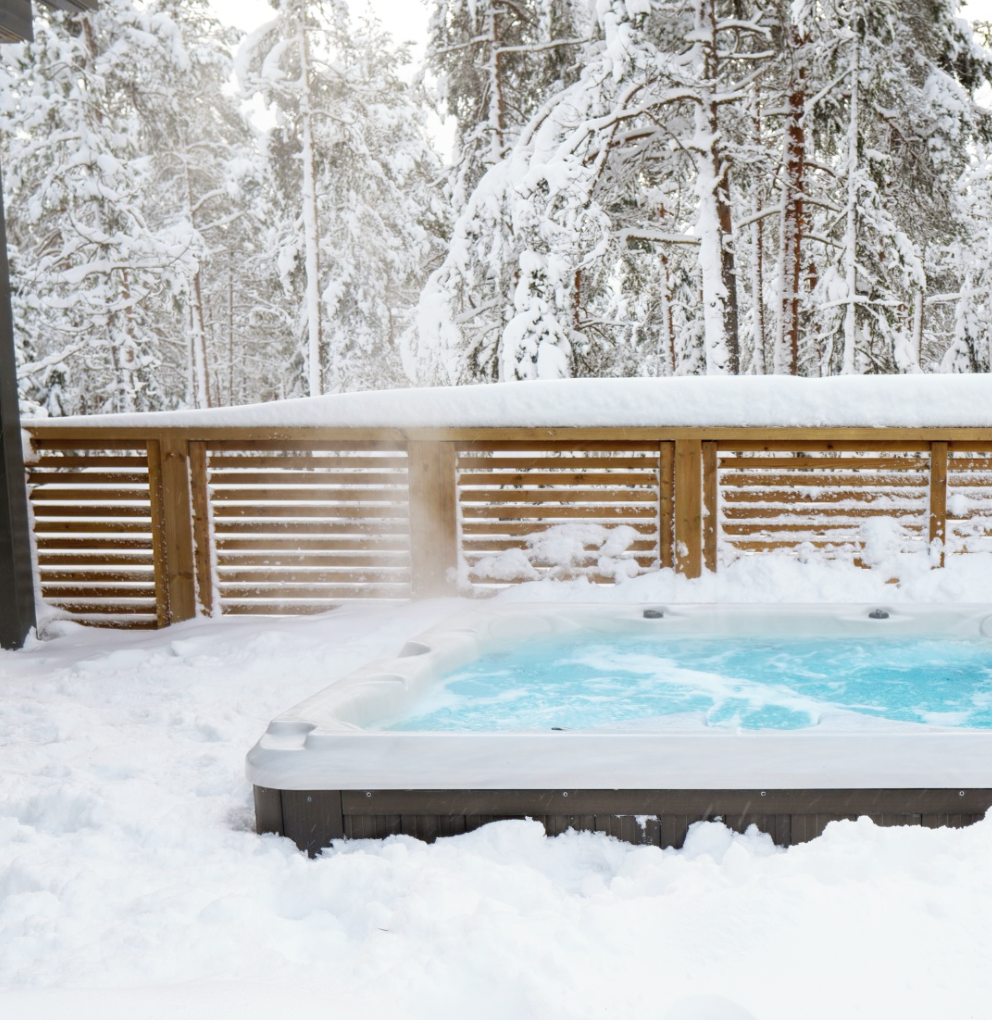 Snow-covered backyard hot tub with steam rising, surrounded by a wooden fence and snow-covered trees in the background.
