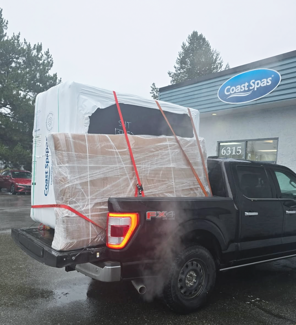 A black pickup truck carrying a large wrapped hot tub in its truck bed, parked outside a Coast Spas store on a rainy day.