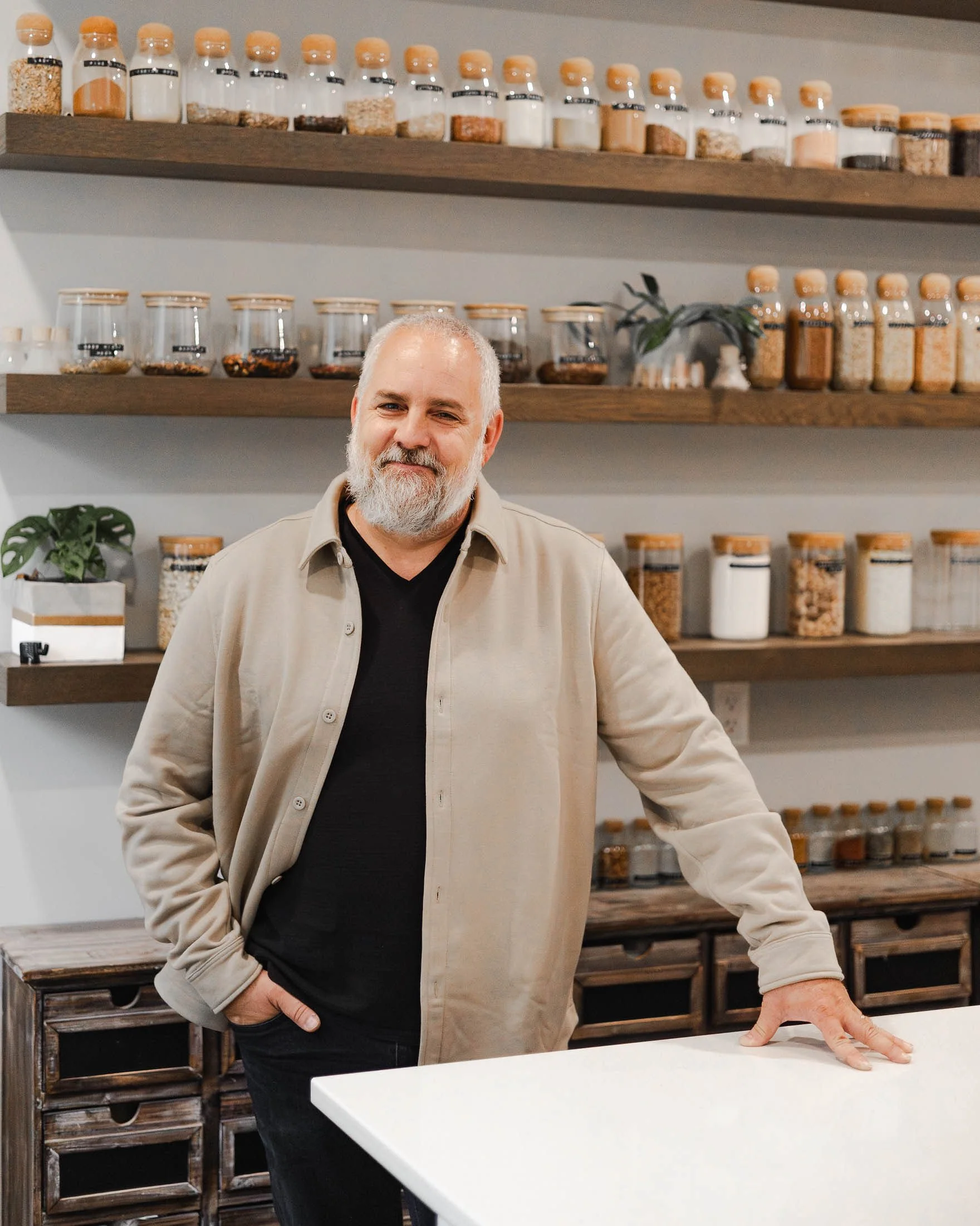 A smiling man with a beard standing in a kitchen or spice shop, leaning on a white counter with his right hand, with shelves of jars filled with spices or grains behind him.