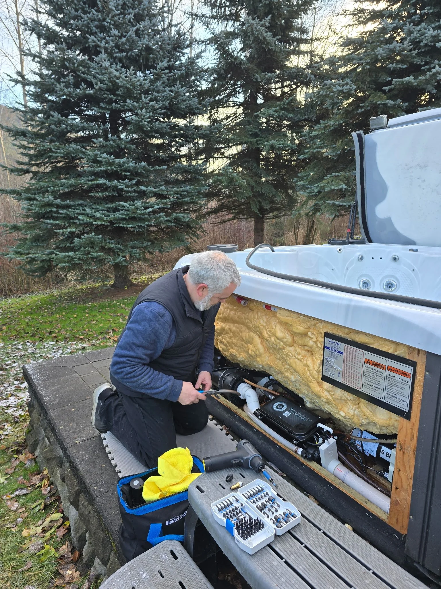 A man kneeling outside next to an outdoor spa hot tub with its side panel removed. He is working on the spa's internal plumbing or electrical components. Tools and a toolbox are placed on a nearby bench, and there are pine trees and some snow on the ground in the background.
