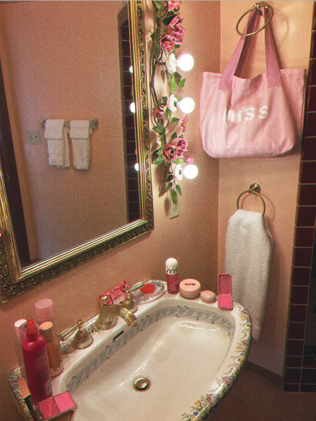 Decorative bathroom sink area with pink and white accents, a mirror, towels, a pink tote bag hanging on a hook that says "Miss" in white with various toiletries on the sink.