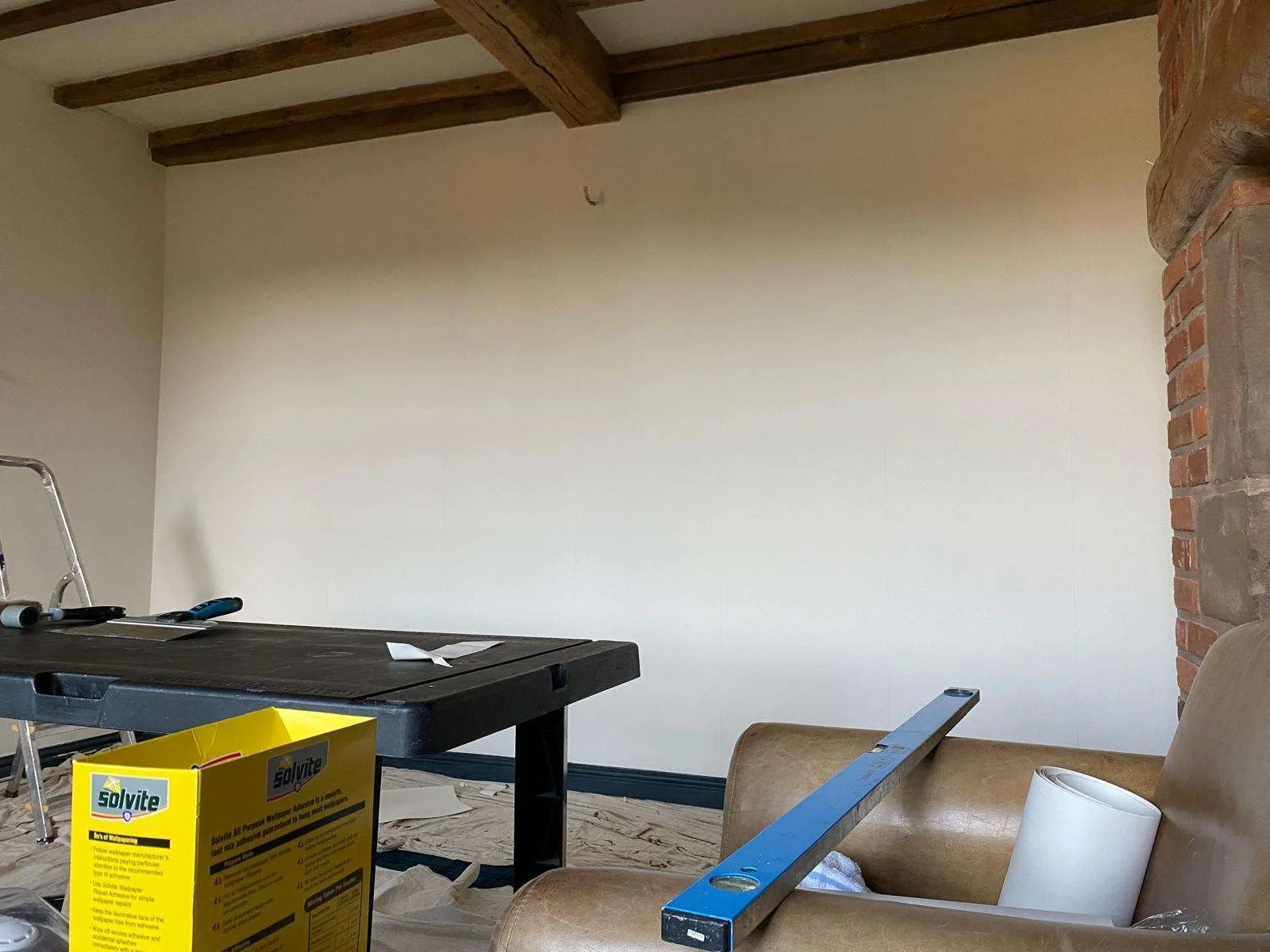 Interior room under renovation with tools and materials scattered; partially visible ladder, a yellow box of Solvite wallpaper paste, a level, a piece of hardware, and a large roll of paper on a beige couch. Exposed brick wall, wooden ceiling beams, and a blank wall.