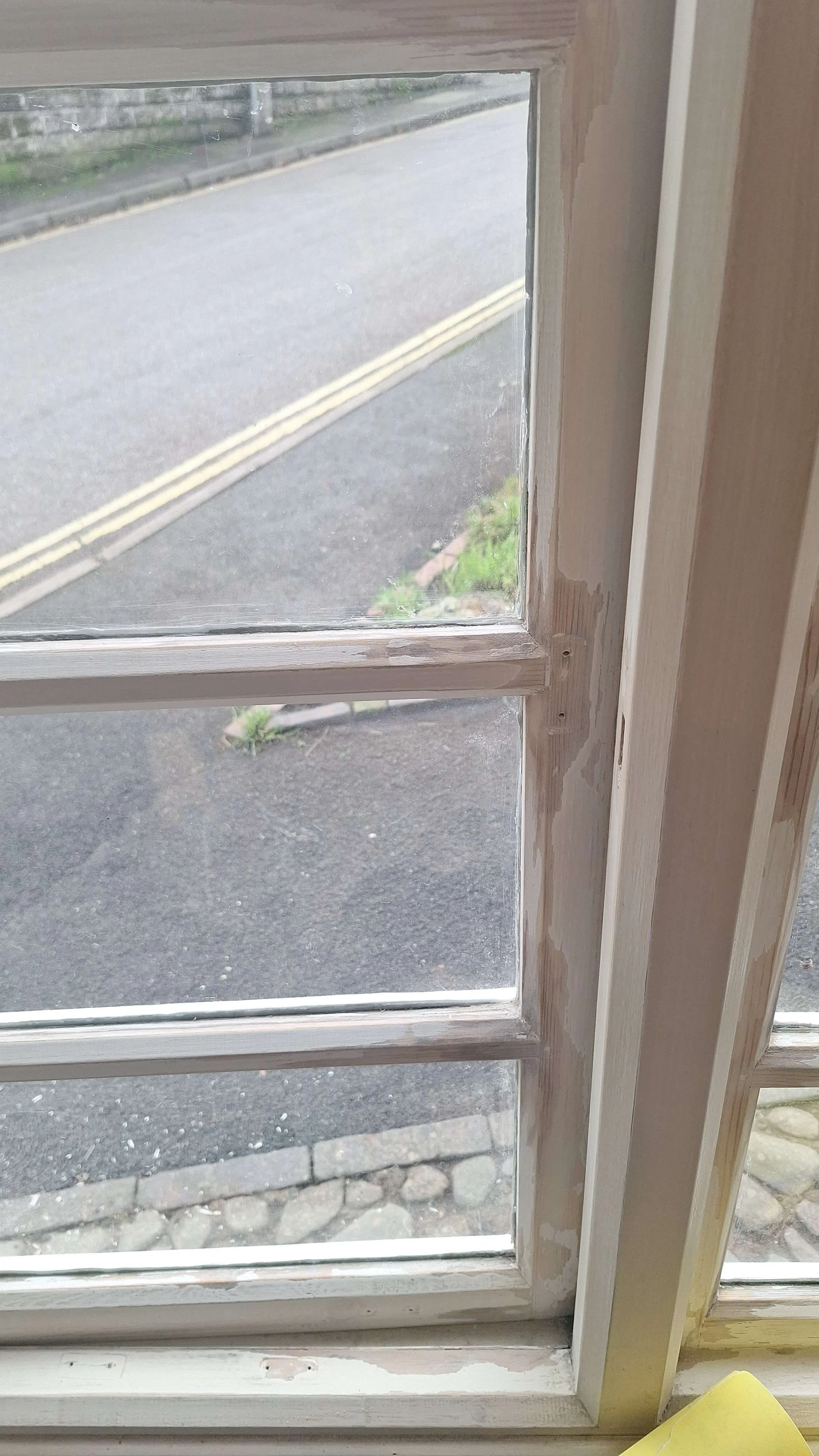 Close-up of a window with a white wooden frame, partially painted, looking out onto a road with a double yellow line and sidewalk.