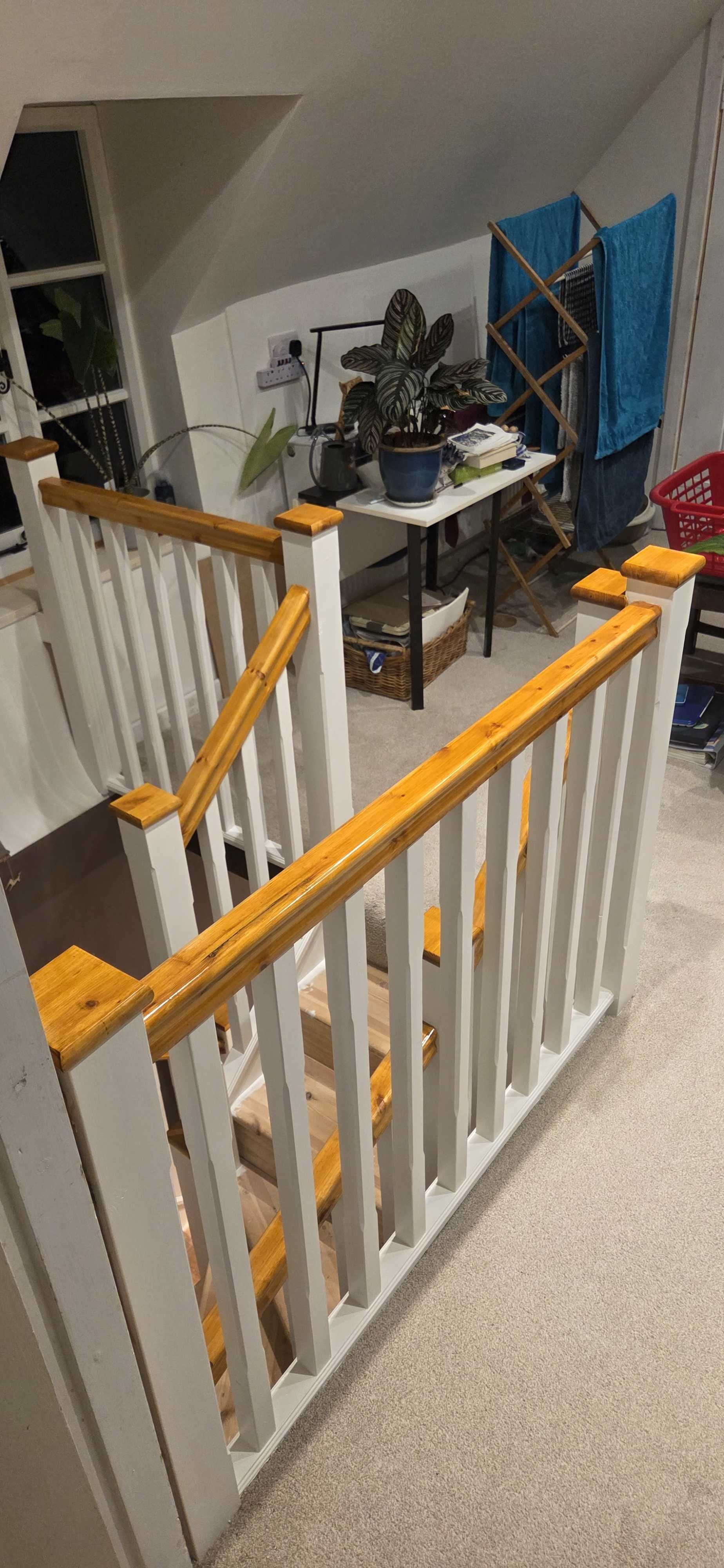 A staircase with white balusters and natural wood handrails leading up to a small landing. In the background, there is a white table with plants, books, and a basket underneath. A folding drying rack with blue towels is visible, along with a window, some plants, and various household items.