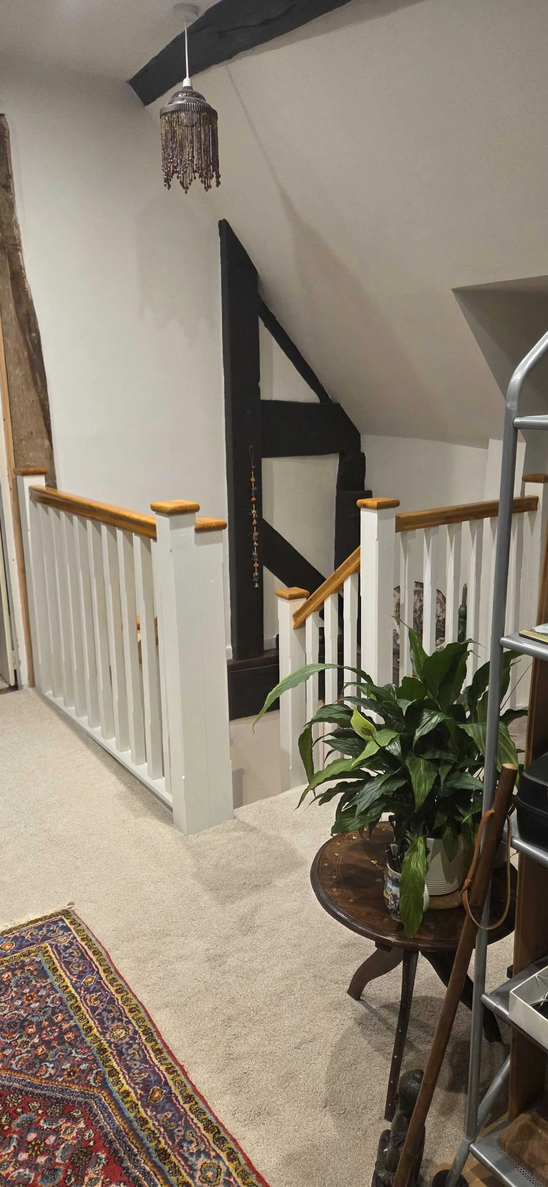 Indoor staircase landing area with white railing and wooden handrails, a large green potted plant on a small wooden table, a colorful patterned rug, a black beam and a black decorative element hanging from the ceiling, a gray ladder on the right, and part of a wall with pictures or artwork.