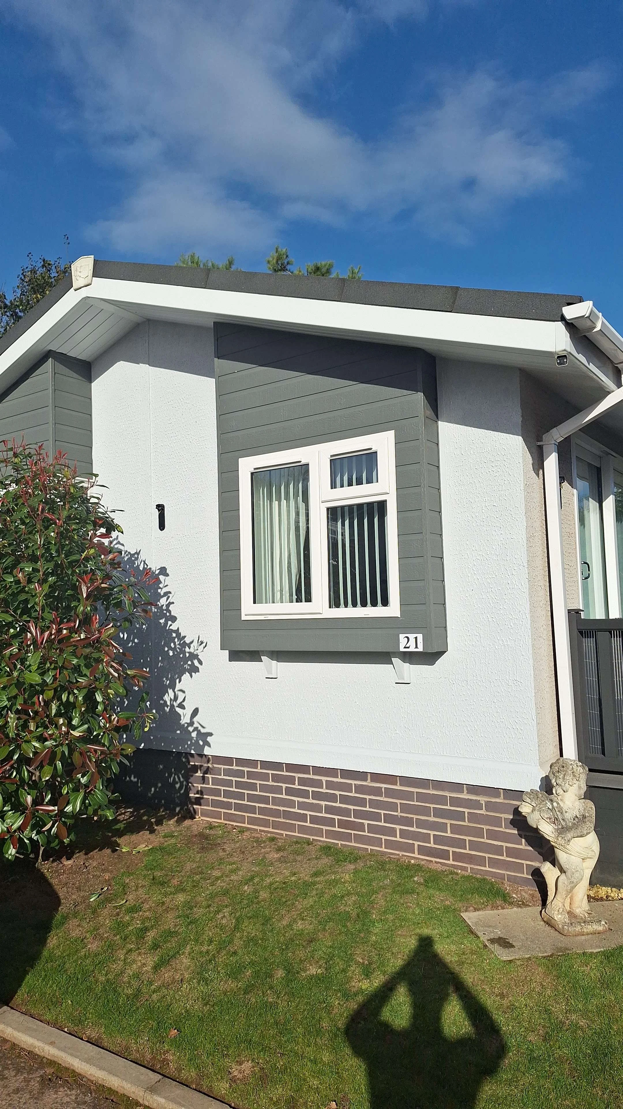 Front view of a modern house with gray and white exterior, a window with blinds, a small garden with grass and a bush, and a decorative stone sculpture near the house.