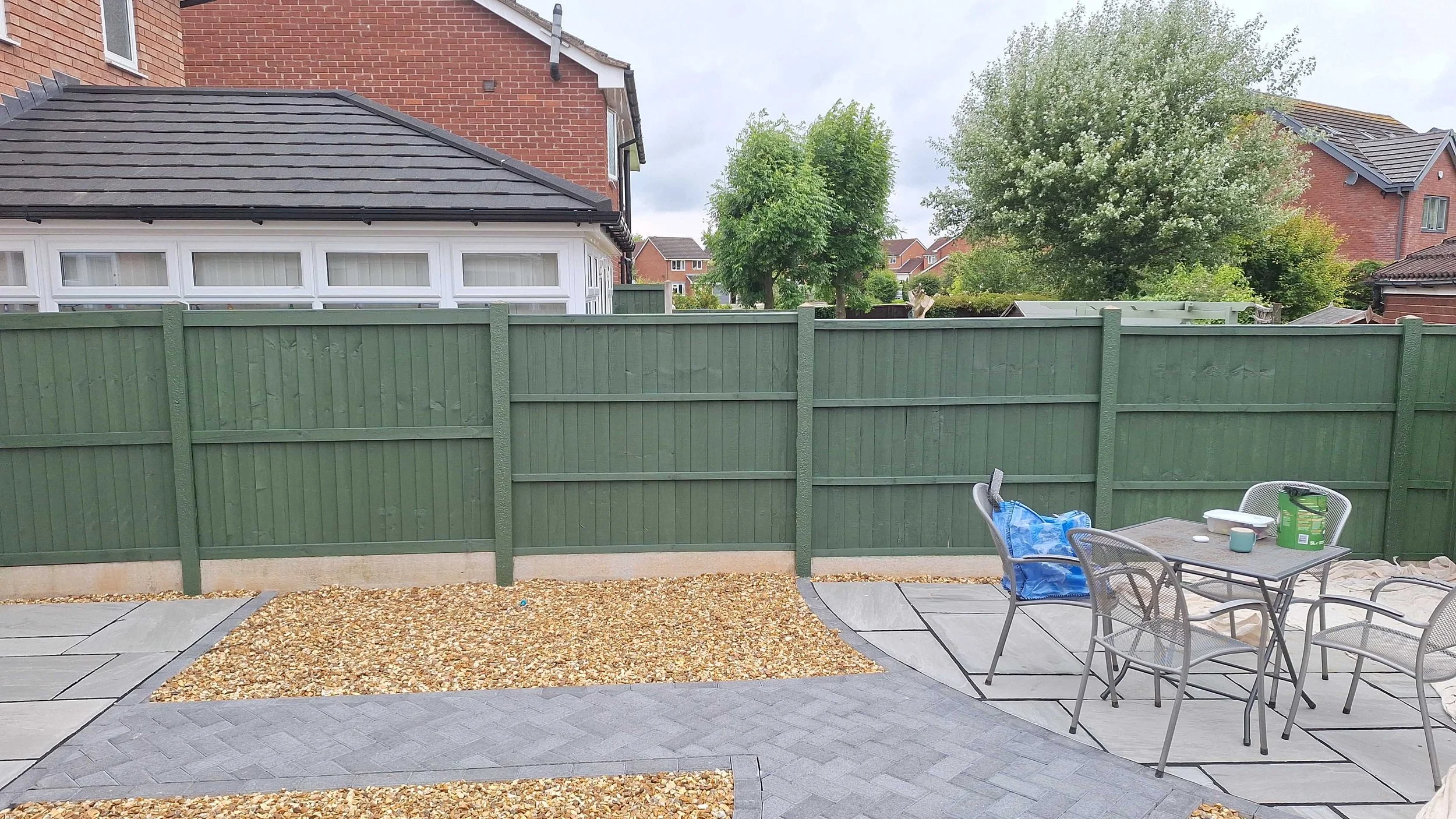 Backyard patio with outdoor table and chairs, green wooden fence, trees, and neighboring houses.