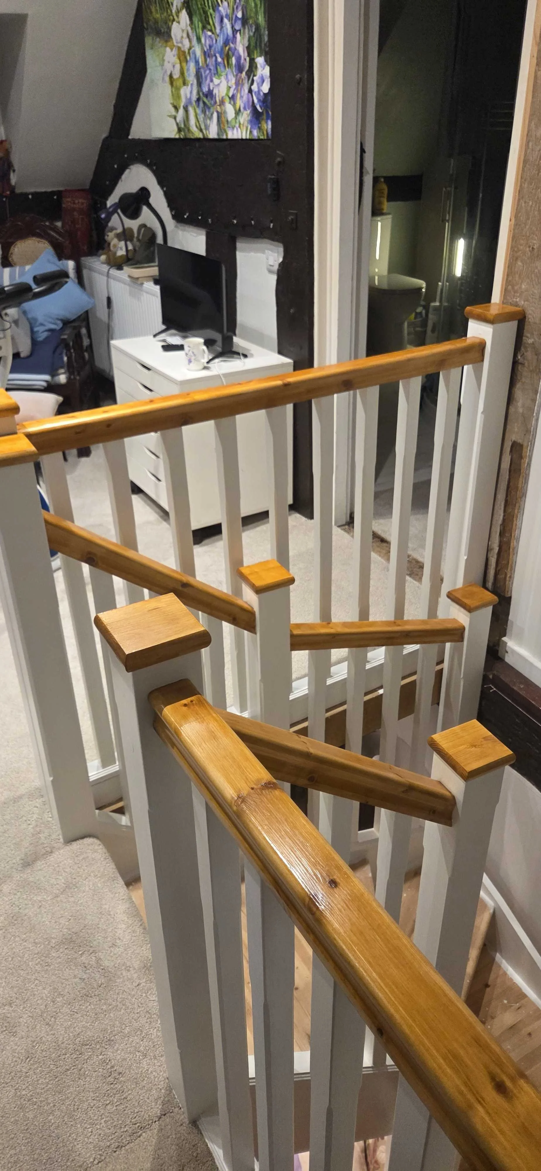 View of a staircase with white railing and polished wooden handrails, leading down to a room with a white cabinet, a small black monitor, and a desk lamp. A colorful floral painting is on the wall, and a bathroom is visible through an open door in the background.