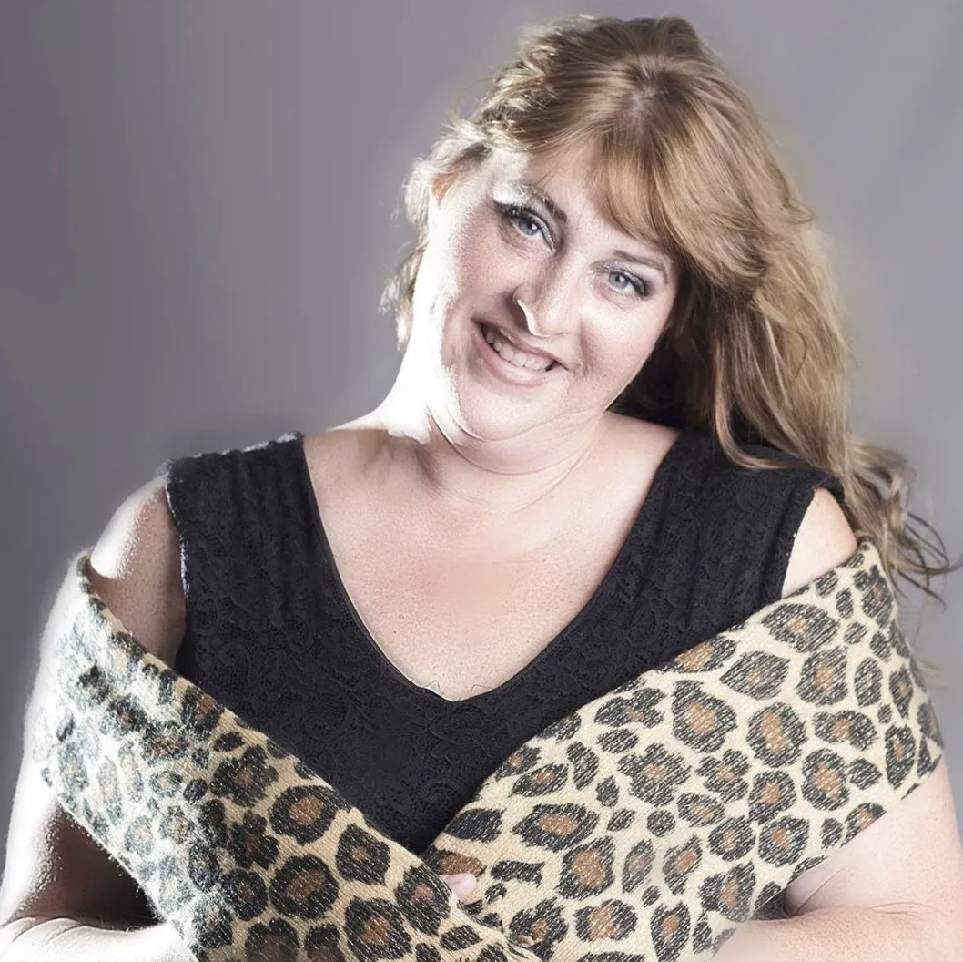 A woman with long reddish hair smiling, wearing a black top and a leopard print shawl.