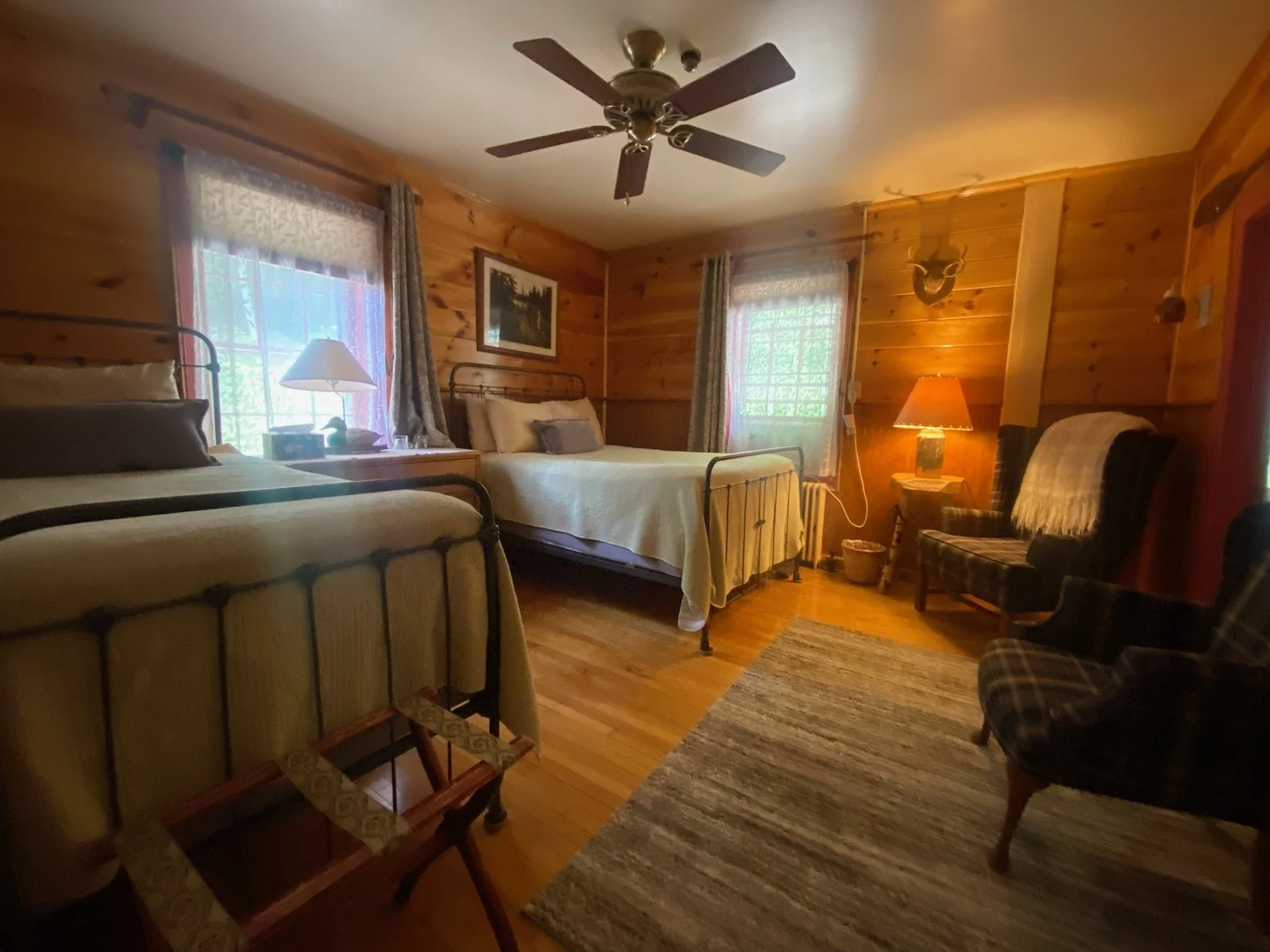 Room with two beds, wooden walls, ceiling fan, window with curtains, armchair, dresser with lamp, and a rug.
