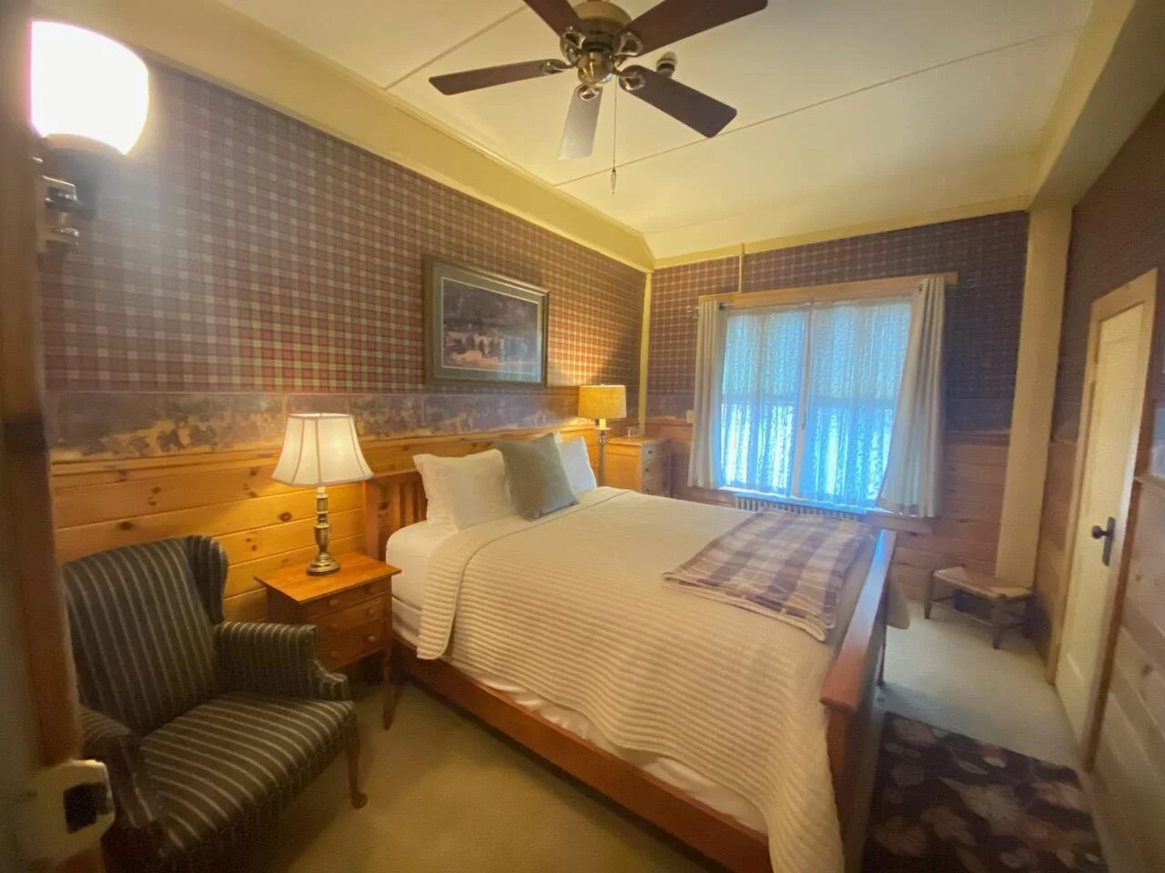 A cozy bedroom with a bed, nightstand, lamps, an armchair, window with curtains, and wooden walls with a plaid wallpaper.