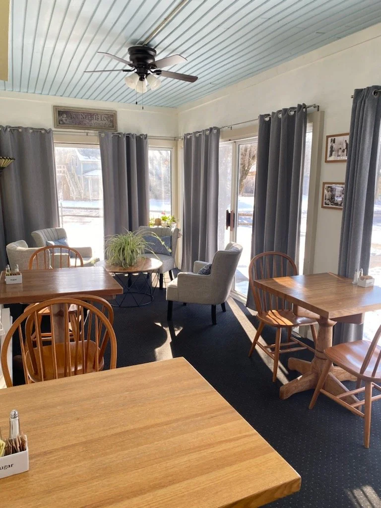 Cozy dining area with wooden tables, wooden and upholstered chairs, gray curtains, a ceiling fan, and a black carpet, with sunlight streaming through large windows.