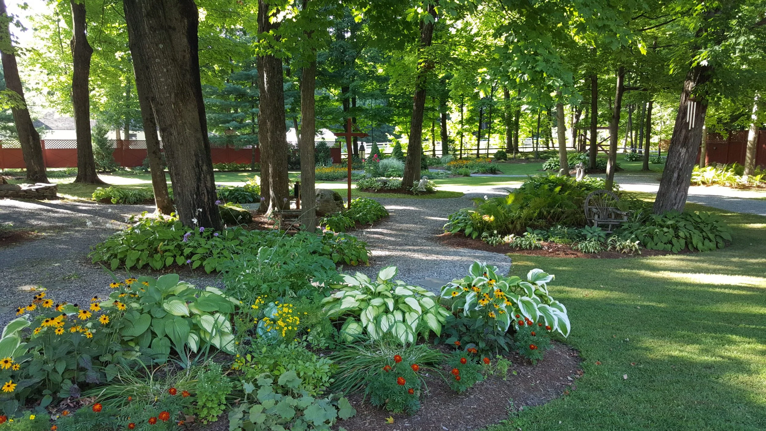A lush garden with tall trees offering shade, surrounded by vibrant flowers, greenery, and a gravel pathway. There are garden ornaments and a wooden bench among the plants.