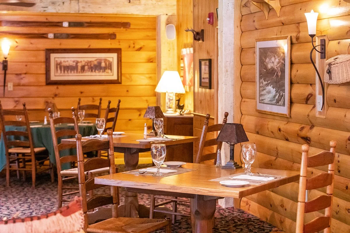 Interior of a cozy rustic restaurant with wooden walls, tables set with glasses and plates, and warm lighting from lamps and wall sconces.