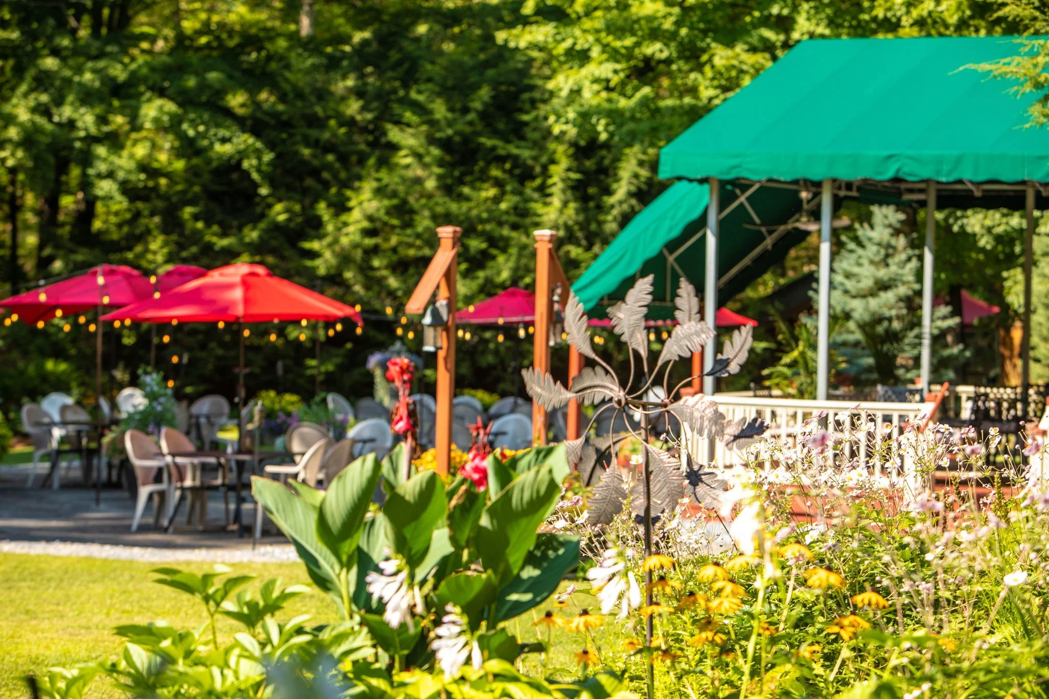 Outdoor garden scene with tables, chairs, and umbrellas, surrounded by lush green trees and flowers.