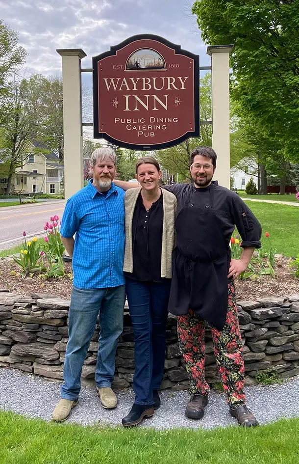 Three people standing in front of a sign for Waybury Inn, with trees and houses in the background. The person on the left is wearing a blue shirt and jeans, the woman in the middle is wearing a black shirt and blue pants, and the person on the right is dressed in chef's attire with floral pants.