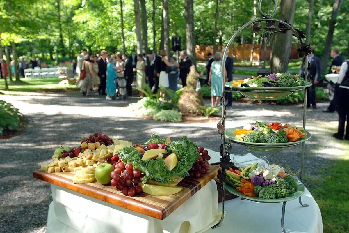 Outdoor gathering with a cheese, fruit, and vegetable platter in the foreground, and people mingling in the background among trees and greenery.