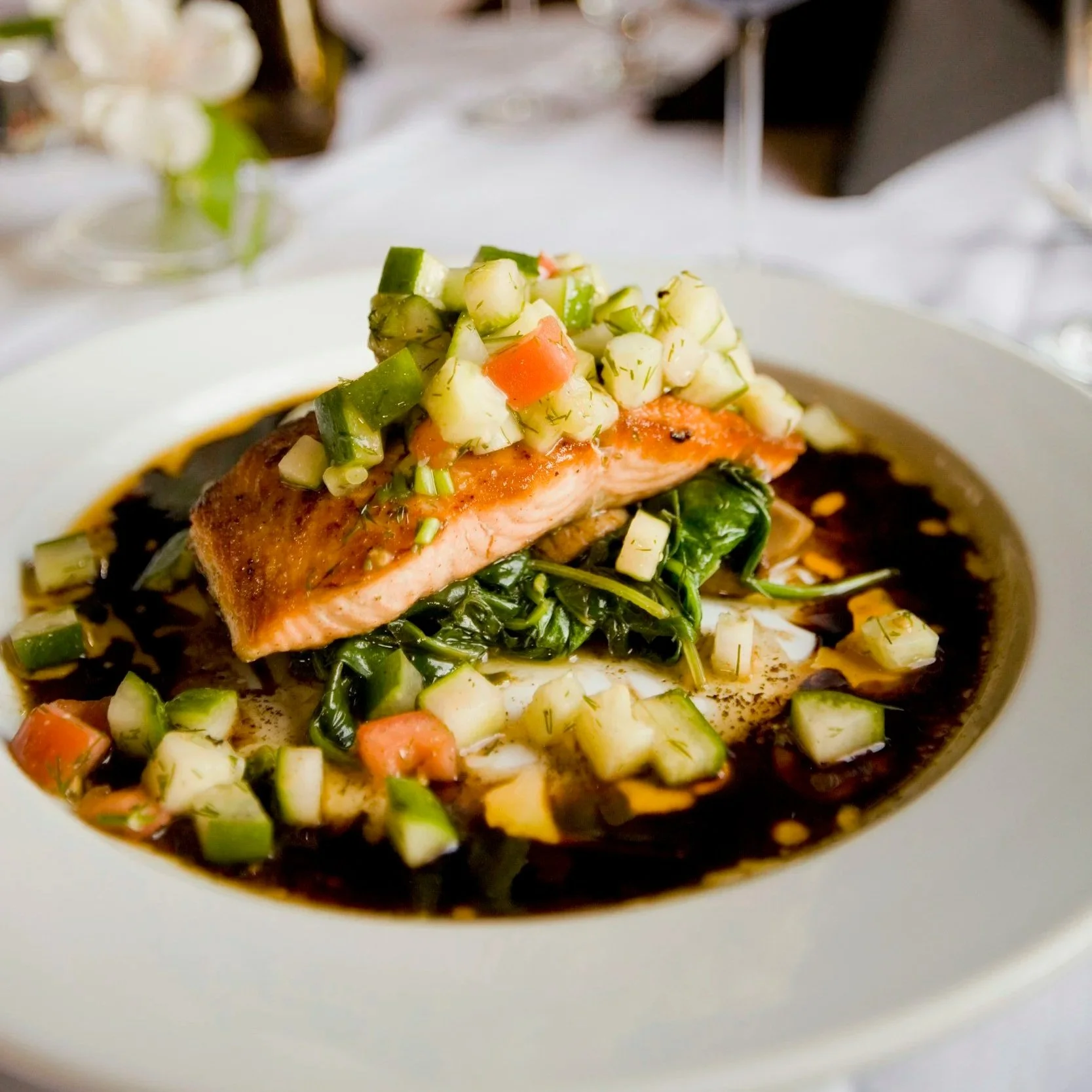 A plate with a cooked salmon fillet topped with diced cucumber, tomato, and herbs, served over greens with a dark sauce or broth.