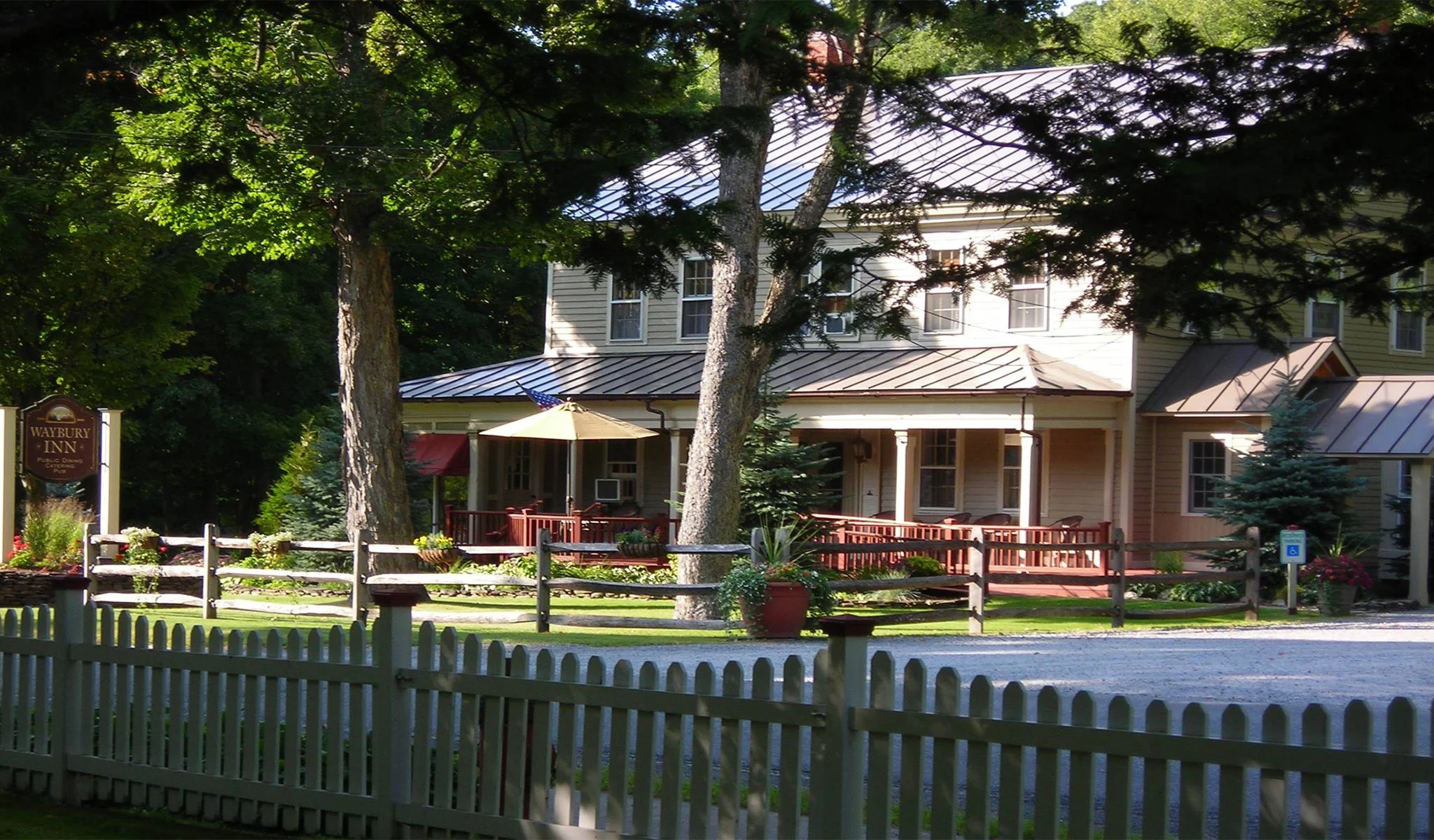 A large two-story guesthouse or inn with a porch surrounded by trees and a white picket fence. The porch has a yellow umbrella, potted plants, and outdoor furniture. A sign on the left reads "Waybury Inn".