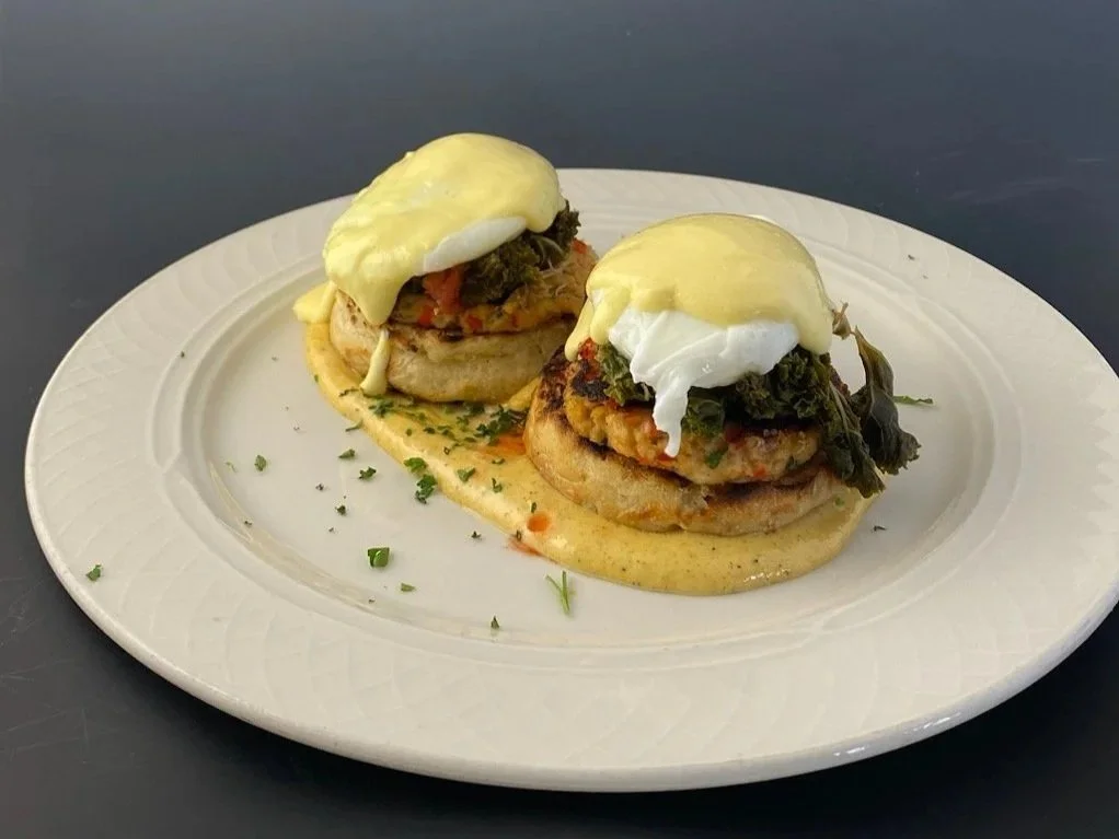Two eggs Benedict dishes on a white plate, topped with hollandaise sauce, served with a creamy yellow sauce on the side, garnished with chopped herbs, and placed on a dark surface.